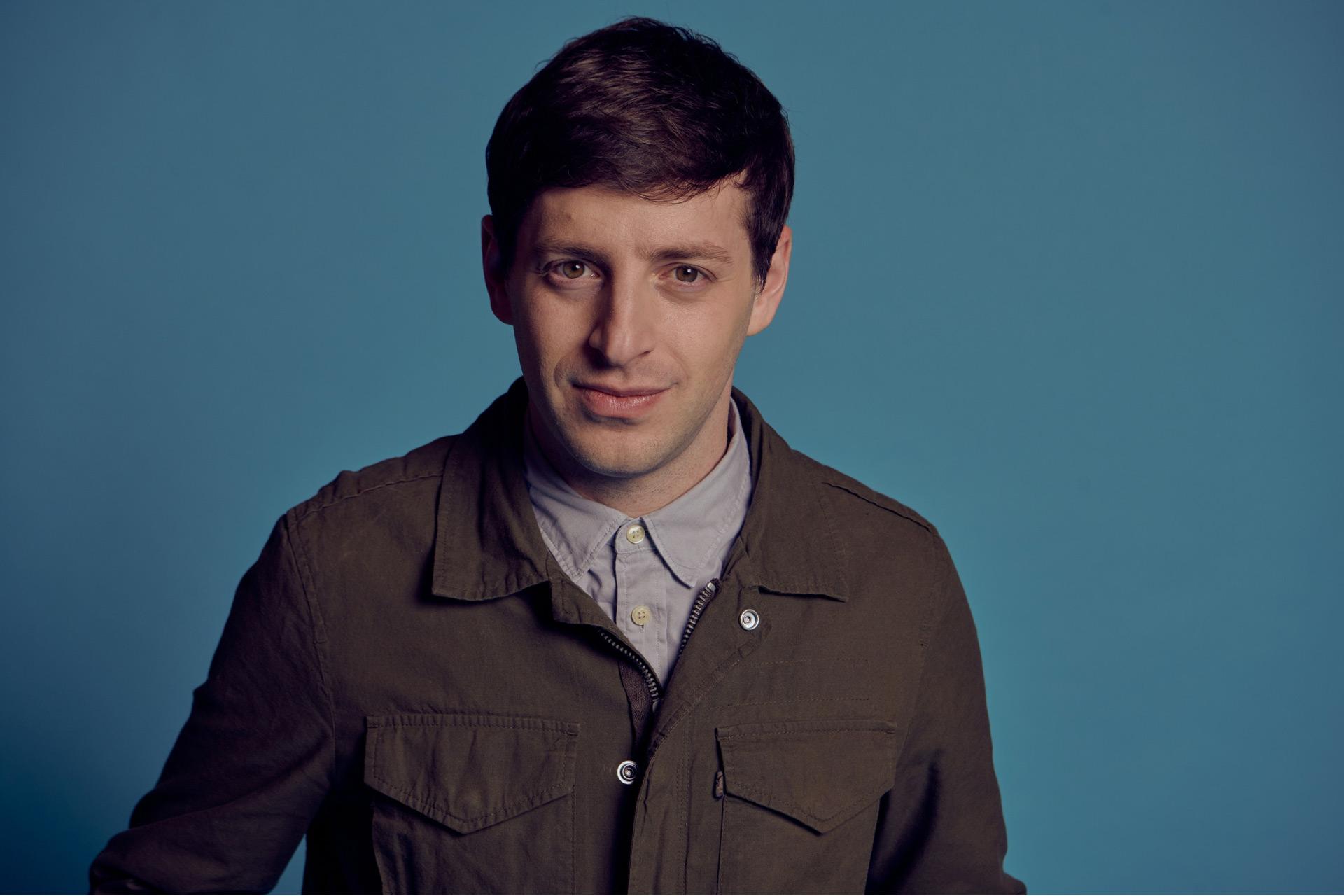 A man with short brown hair wearing a green jacket over a buttoned-up light gray shirt stands in front of a plain blue background, looking directly at the camera with a slight smile.