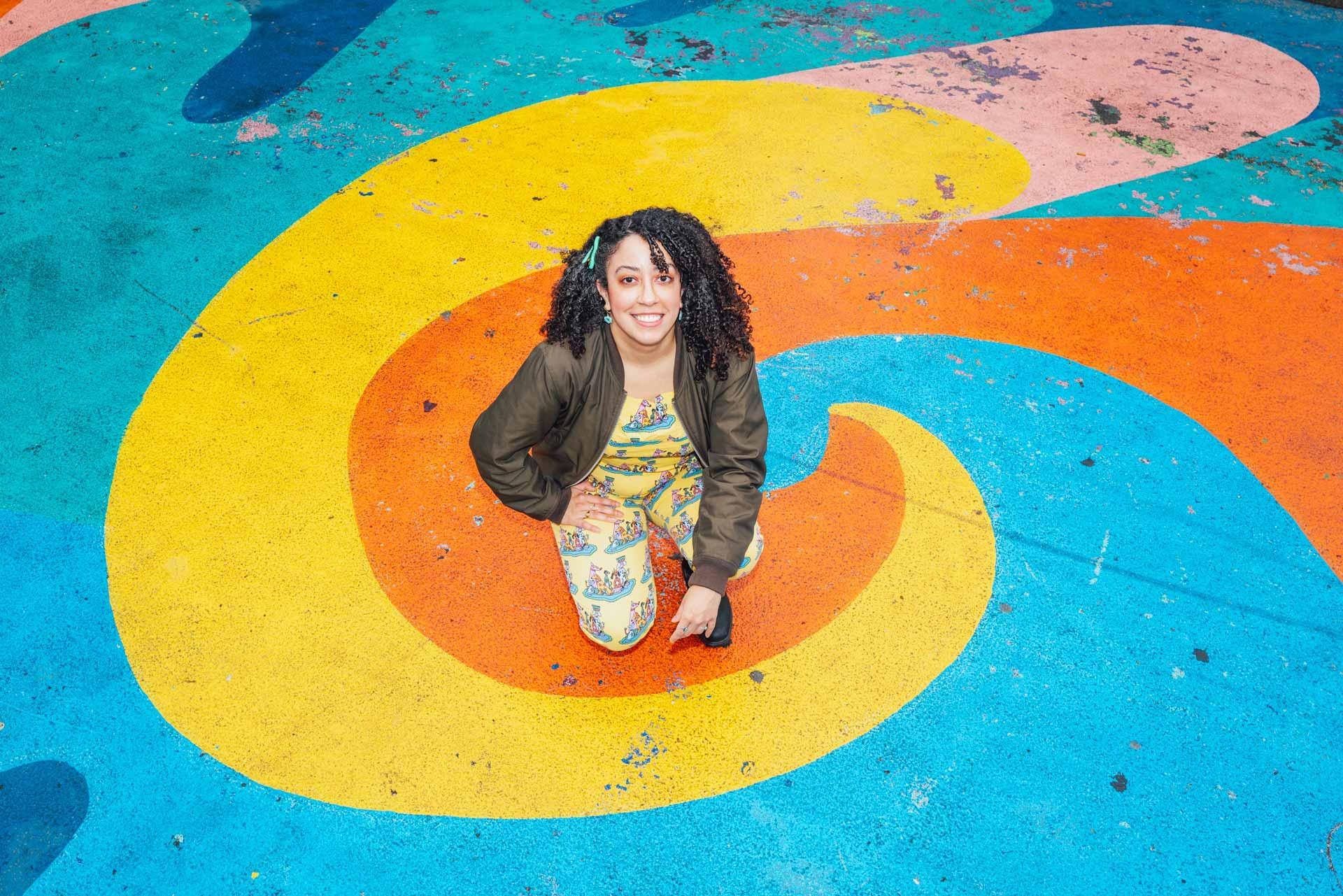Talisa Almonte kneels on a vibrantly painted mural with swirling yellow, orange, pink, and blue patterns. She wears a floral outfit and a dark jacket, smiling up at the camera.