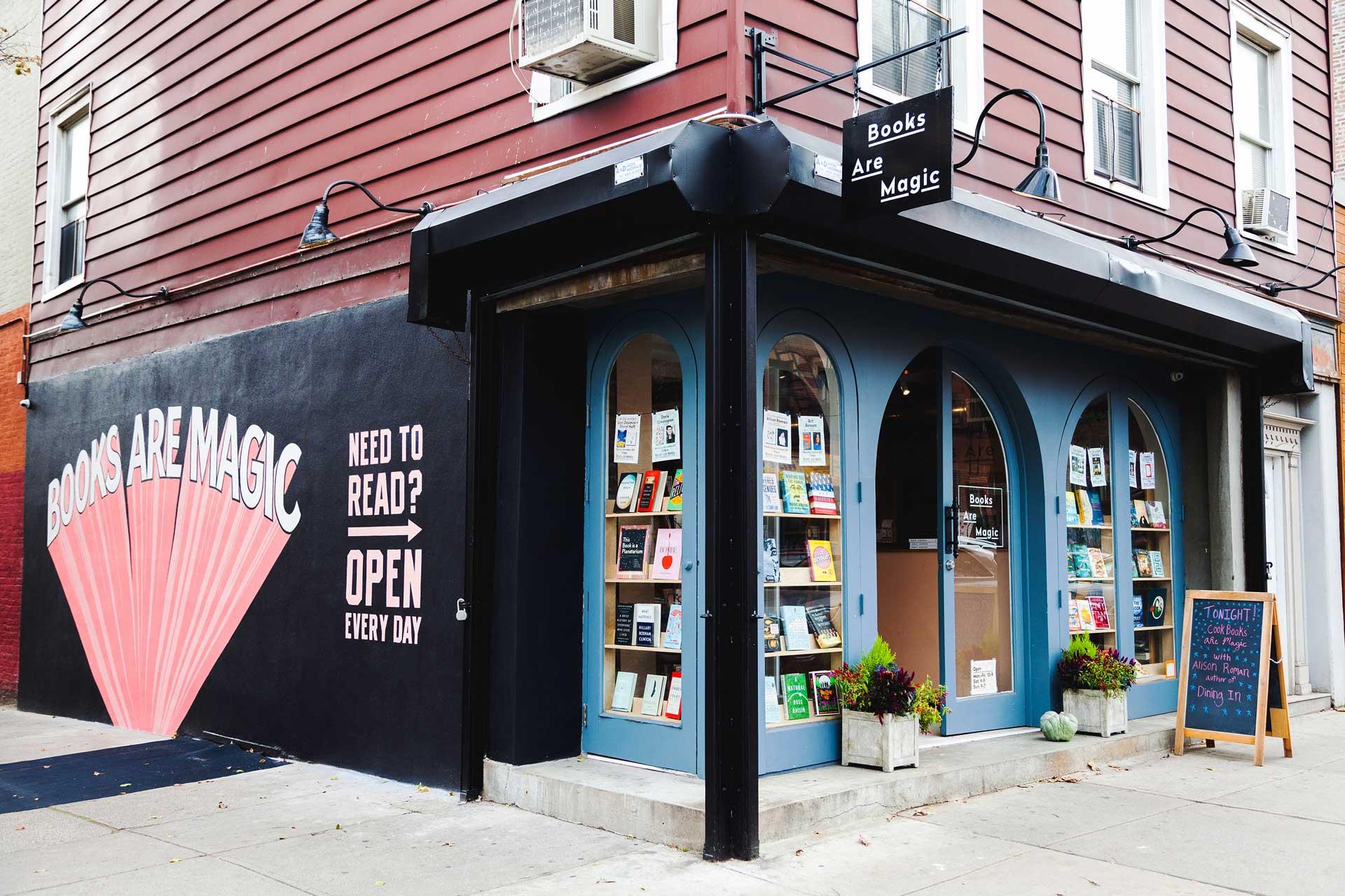 Storefront of a bookstore with curved windows displaying books. The wall reads "Books Are Magic" in large letters and "Need to Read? Open Every Day." The entrance is on a corner with a sandwich board sign outside on the sidewalk.