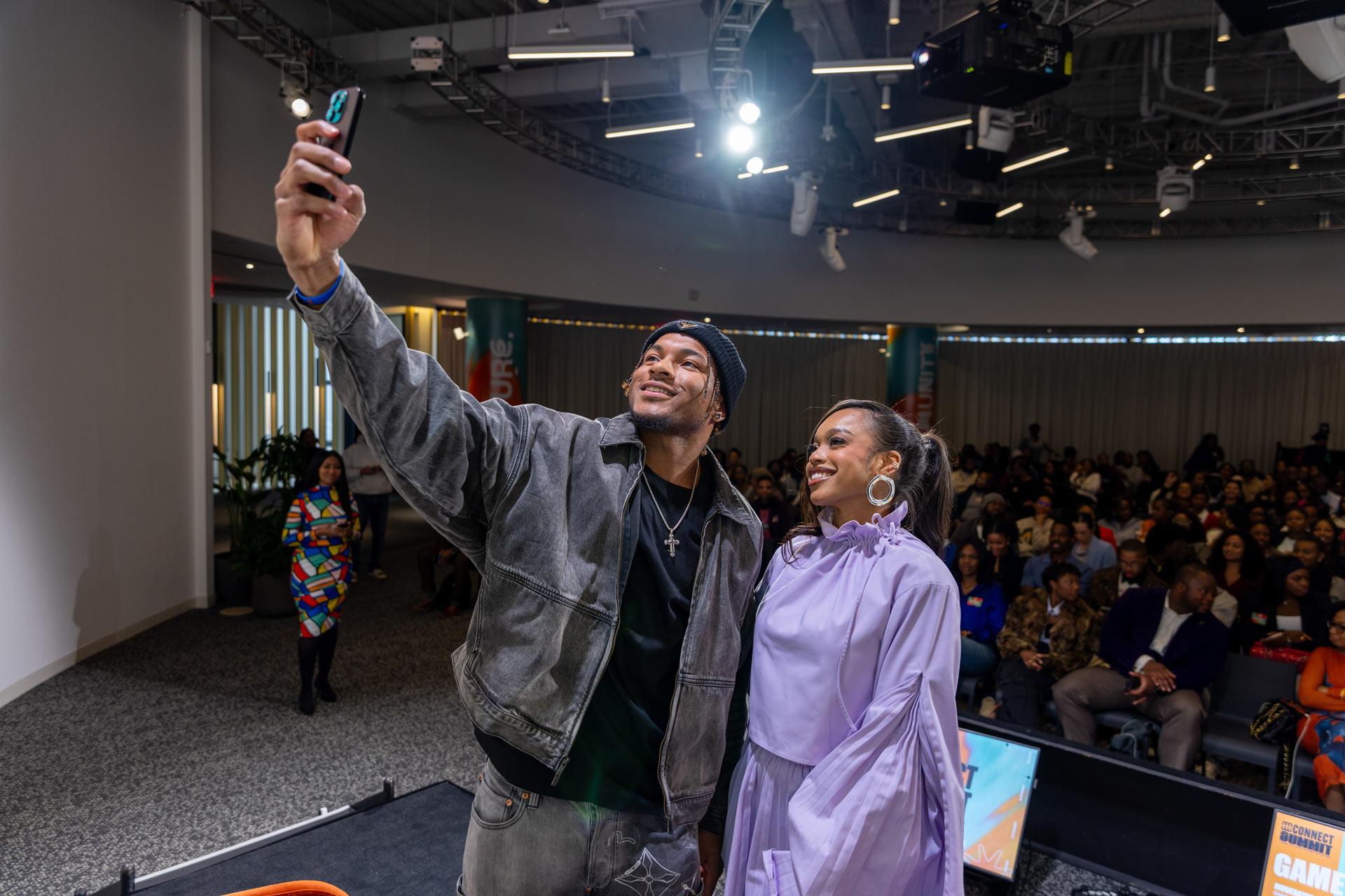 Two people stand on a stage taking a selfie together, smiling at the camera. An audience sits behind them in a modern, well-lit event space with spotlights and a curved ceiling.
