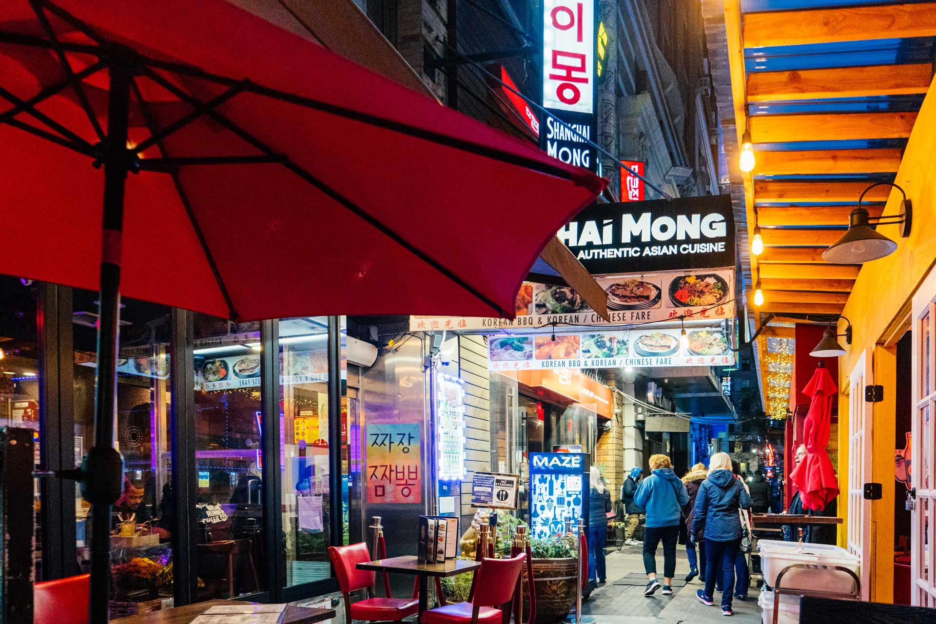 A vibrant, colorful street scene at night shows people walking past restaurants with bright neon signs, outdoor seating, red umbrellas, and various Asian cuisine advertisements lining the narrow alley.