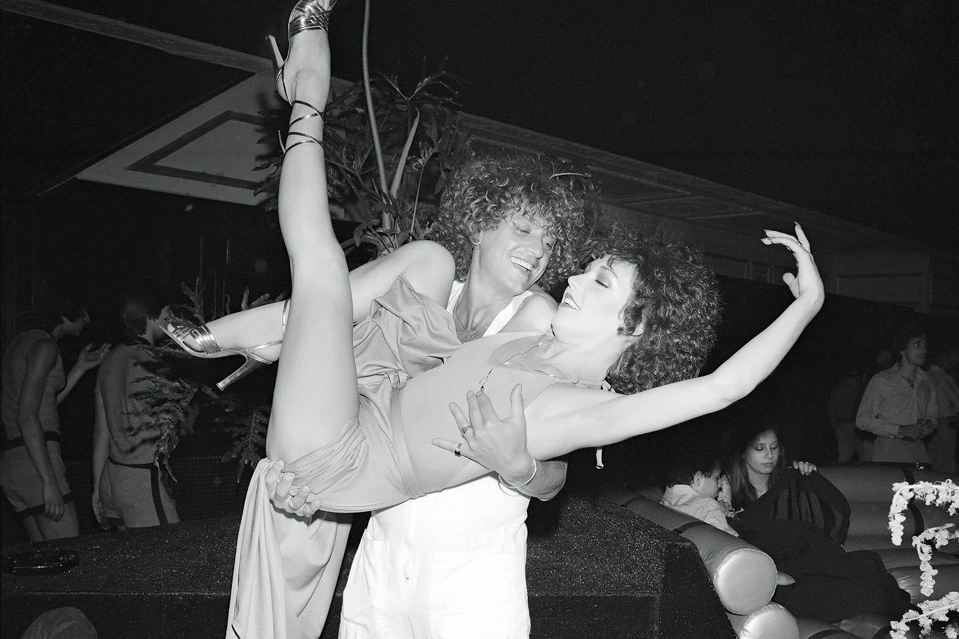 Two people with curly hair joyfully pose together in a lively, dimly lit nightclub. One person lifts the other, who raises a leg high, both smiling and appearing to dance. Other guests relax in the background.