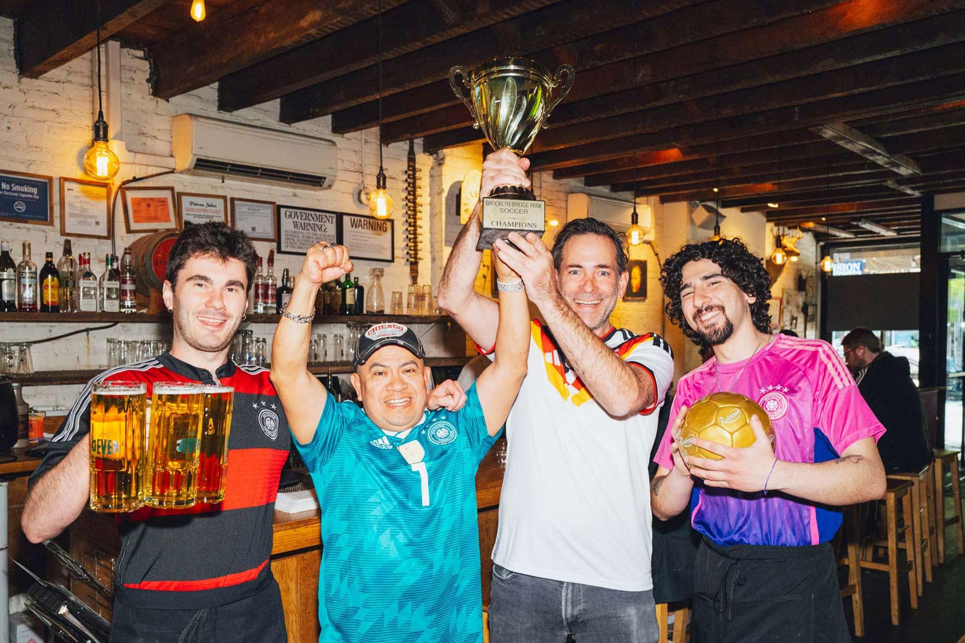 Four smiling people pose in a bar wearing soccer jerseys; one holds two large beer mugs, another raises a fist, the third lifts a trophy, and the fourth holds a gold soccer ball. The bar has wooden beams and hanging lights.