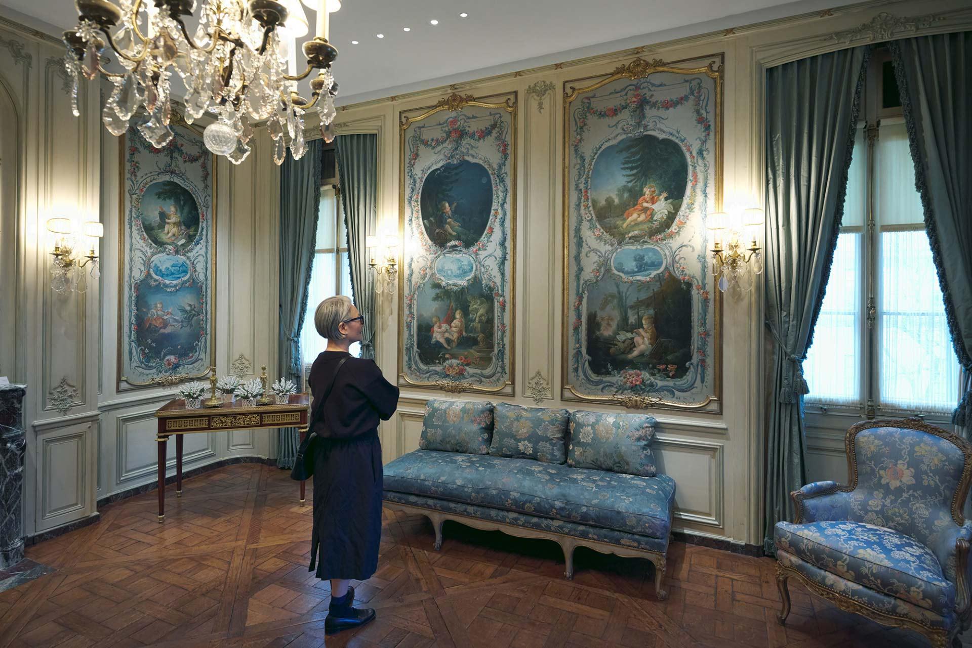 A person with short gray hair stands in an ornate room, viewing large, decorative paintings on the walls. The room has elegant furniture, curtains, wooden floors, and a sparkling chandelier.