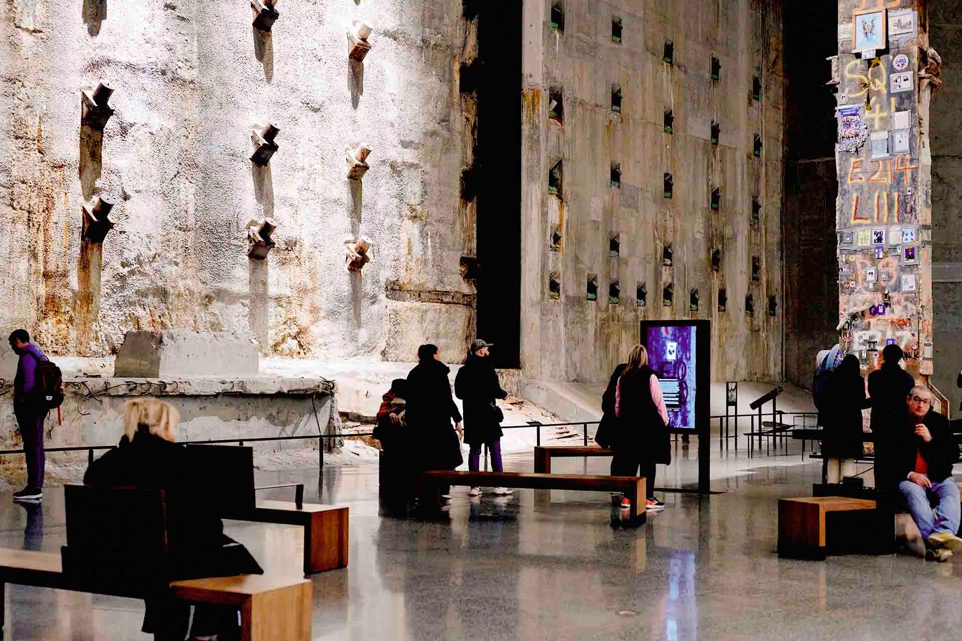 People sit and stand in a large, somber museum space with concrete walls, metal star-shaped anchors, and memorial displays. The atmosphere is quiet and reflective.