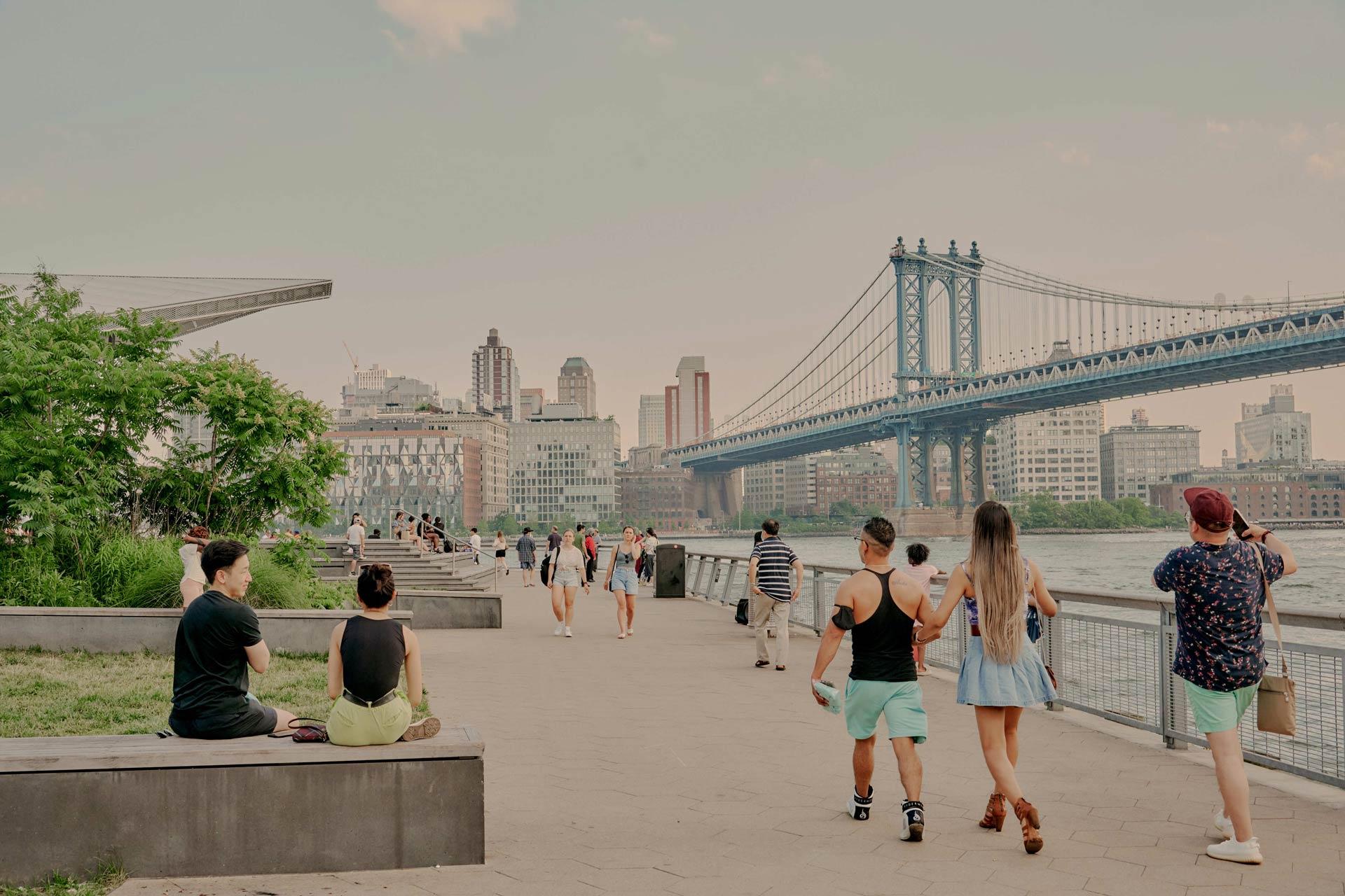 People stroll along a riverside walkway near a large suspension bridge. Some sit on benches, while others walk. Tall buildings are visible in the background under a cloudy sky.