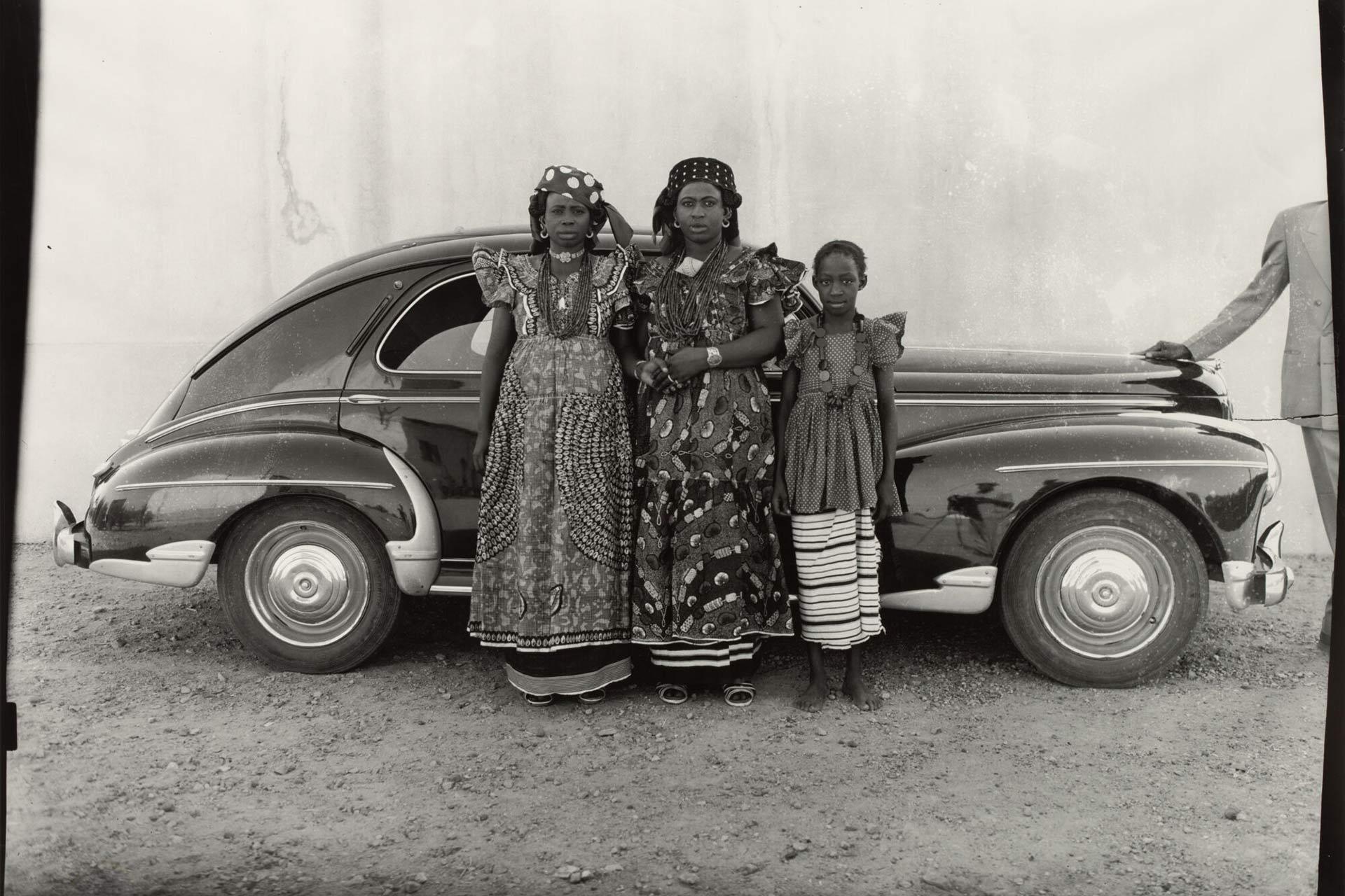Three people in traditional patterned clothing stand in front of a vintage car. Two adults are in ornate dresses and headpieces, while a child stands beside them. The background is a bare wall, and part of another person is visible on the far right.