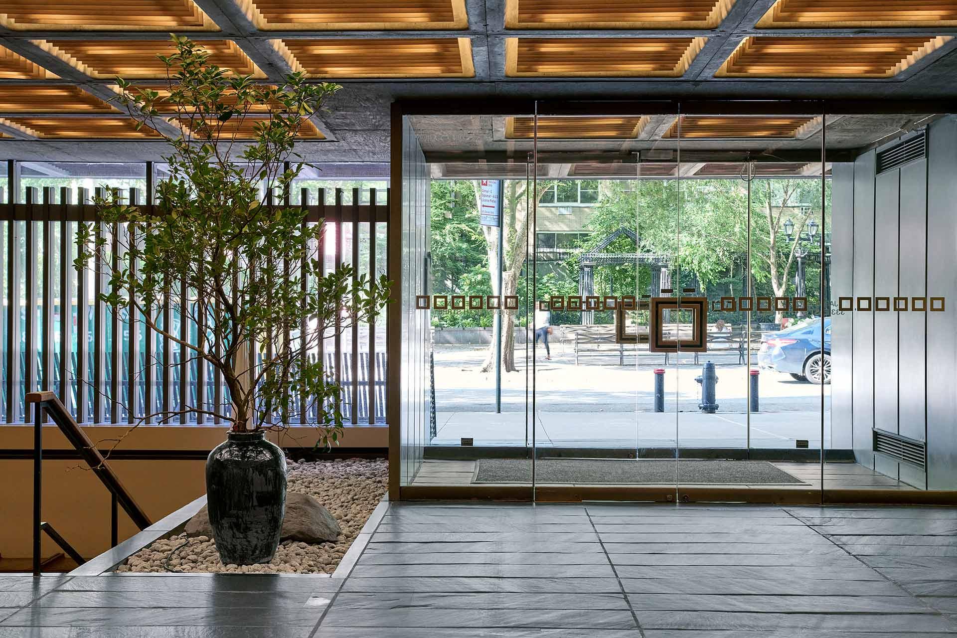 Modern building lobby with large glass doors, a potted tree on a bed of stones, and wood-paneled ceiling. Sunlight and greenery can be seen outside through the glass entrance.