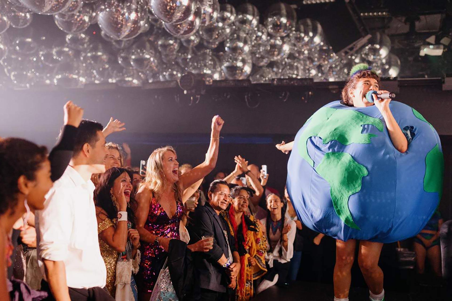 A performer dressed in a large Earth costume sings into a microphone on stage, while a lively crowd cheers and captures the moment under shiny disco balls.