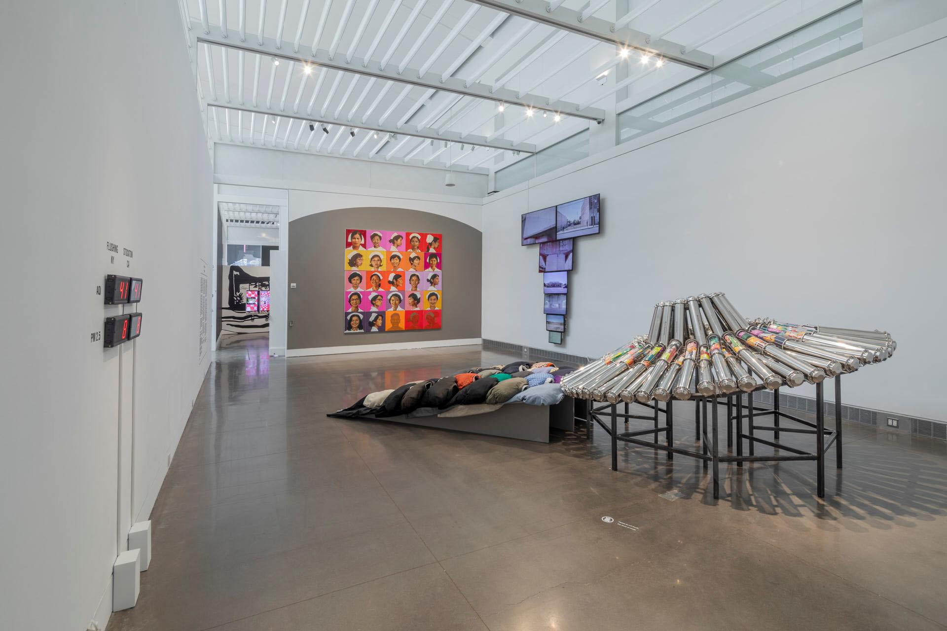 A contemporary art gallery features colorful pop-art portraits, a sculpture of bundled metallic tubes, a pile of pillows with printed faces, and digital displays on a white wall under a bright, industrial ceiling.