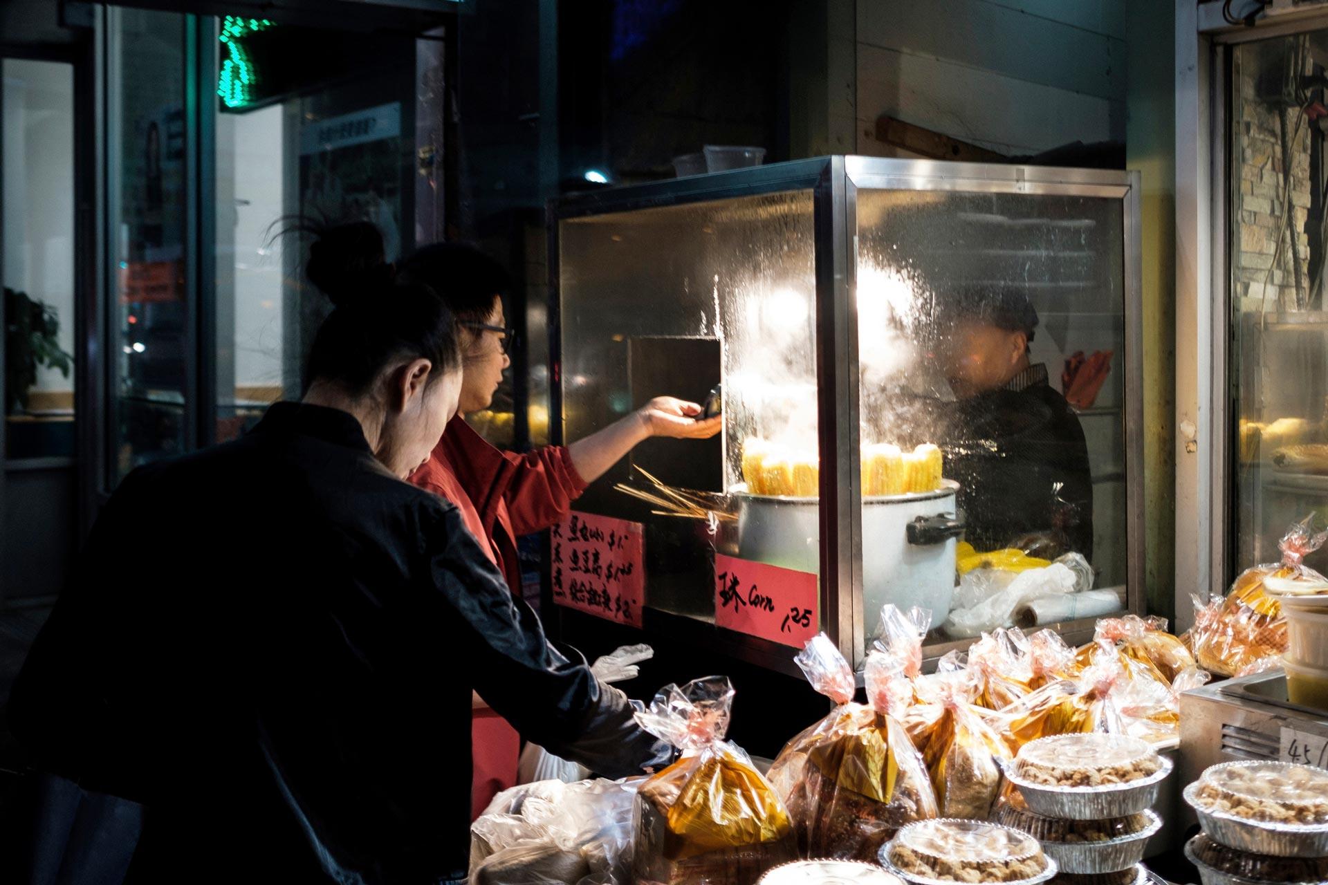 Two people standing at a street food stall at night. A woman in a red jacket serves food from a steaming cart, while a person in black selects items from a table filled with packaged goods. The scene is warmly lit, highlighting the steaming food.