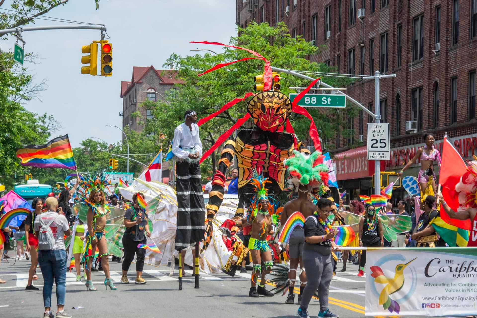 People in vibrant costumes, some on stilts, parade down a city street with colorful flags and banners. A large, elaborate puppet figure is in the center. Spectators line the sidewalk. Buildings and trees are in the background.
