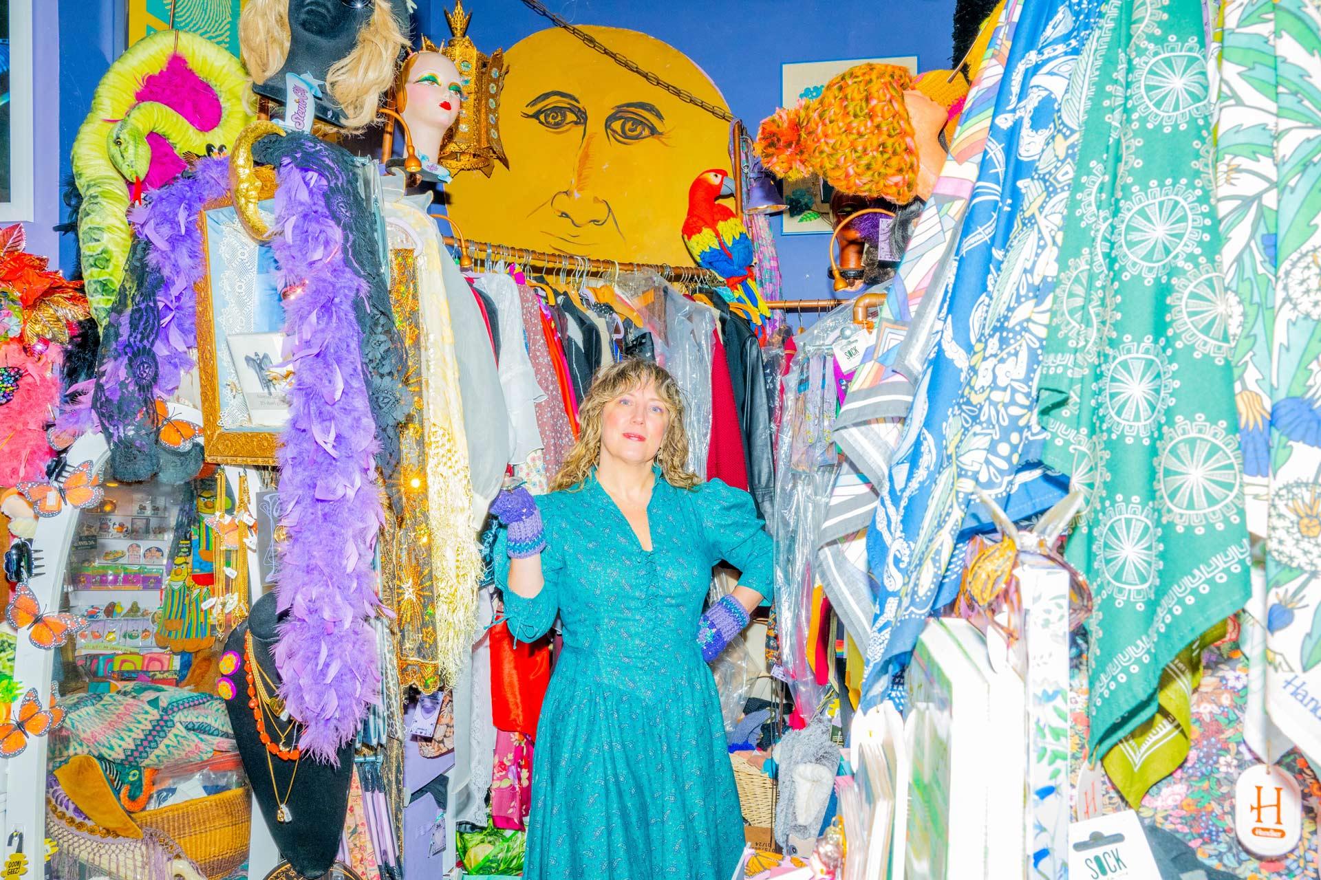 Rebecca Vicars, in a bright teal dress and purple gloves, stands in a colorful, crowded vintage clothing shop filled with patterned fabrics, hats, boas, and a wall with art behind her.