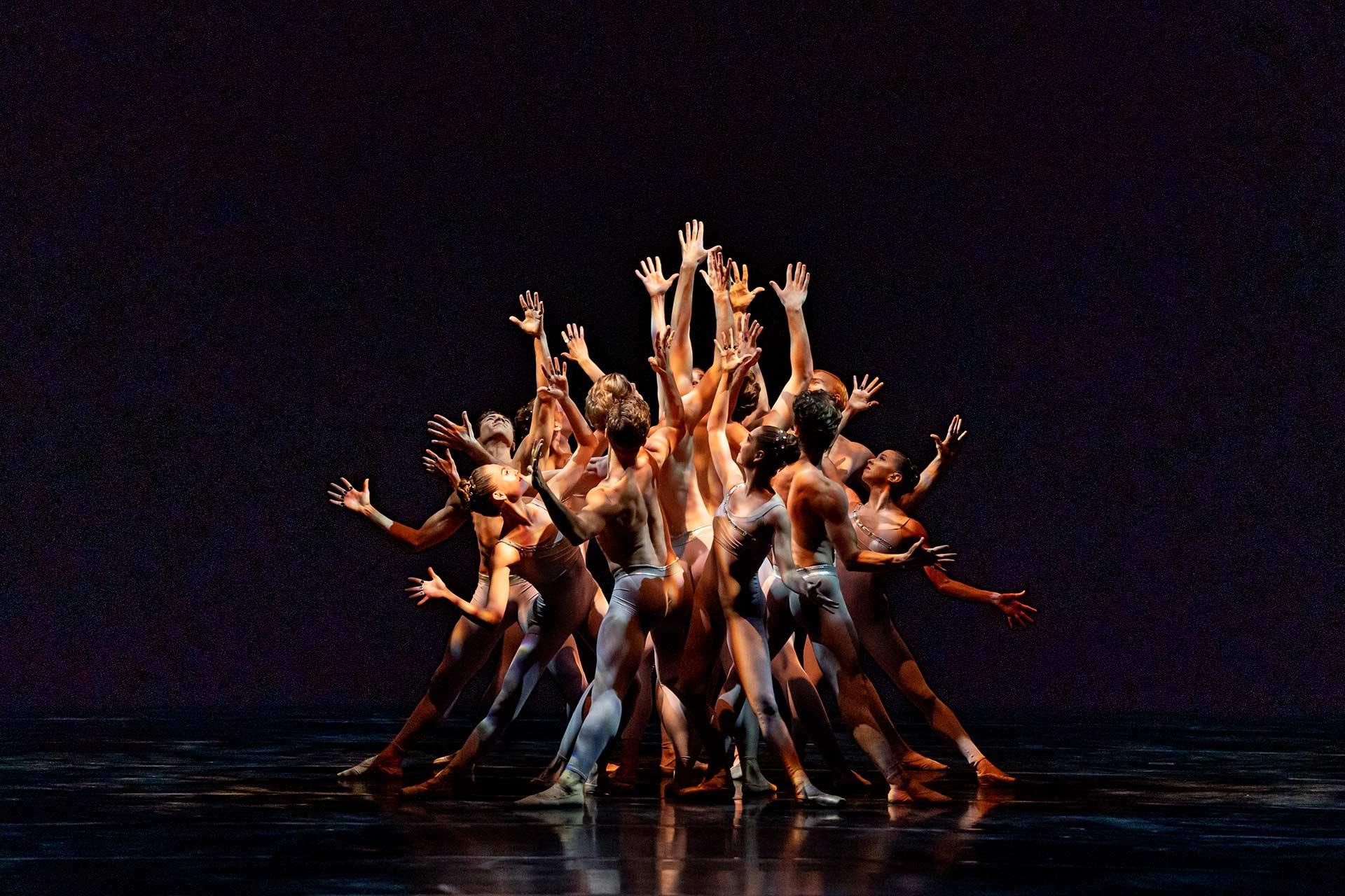 A group of dancers in nude-colored costumes stand close together on stage, reaching their arms upward and outward dramatically against a dark background. Their bodies and movements create an intricate, expressive formation.