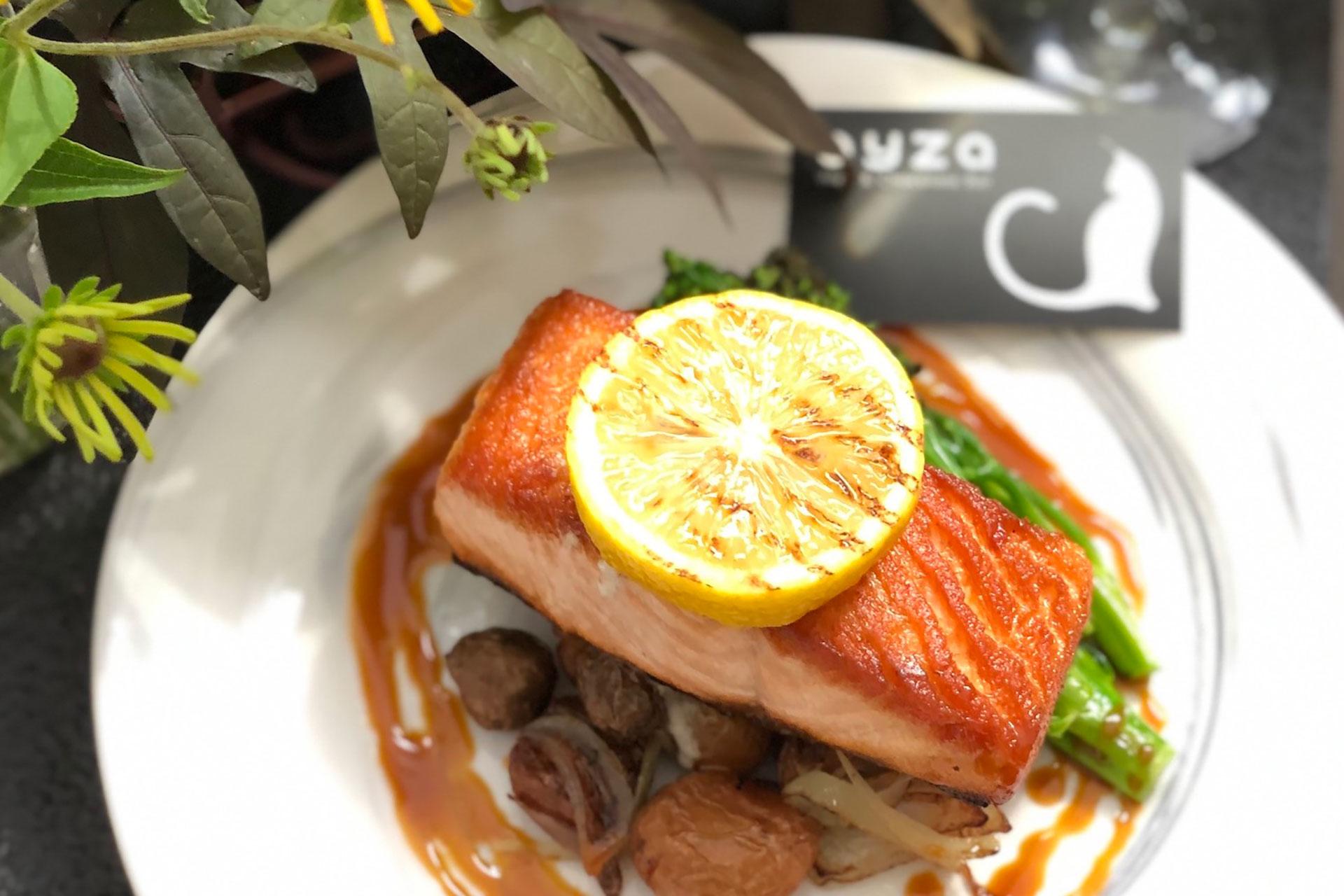 A plate with a seared salmon fillet topped with a lemon slice, served over roasted potatoes and asparagus, garnished with sauce. A flower and a card with a cat logo are nearby on the table.