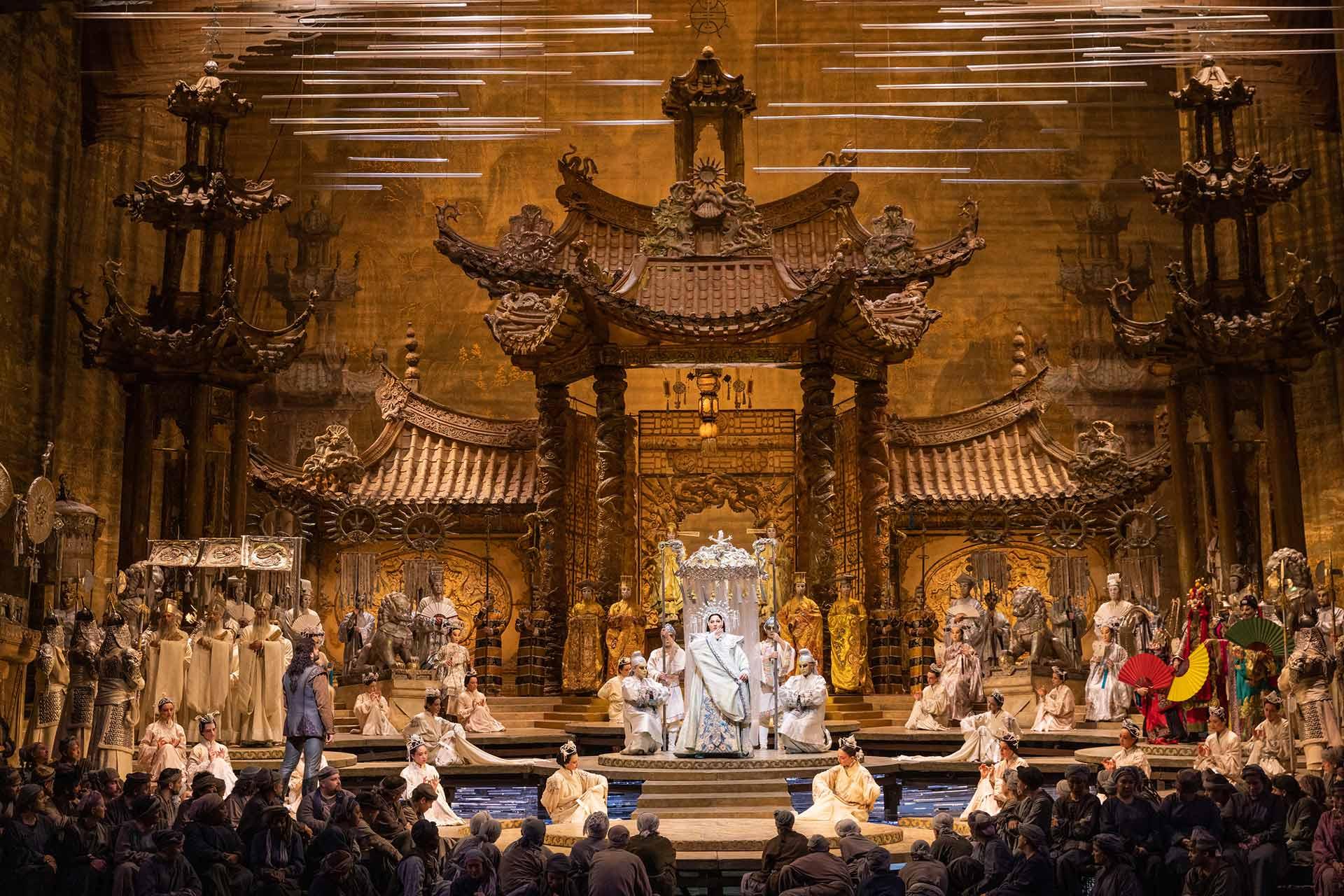 A grand opera stage features ornate, golden, Asian-inspired architecture. Performers in elaborate costumes surround a central figure seated on a throne, while others stand or kneel, creating a dramatic, theatrical scene.