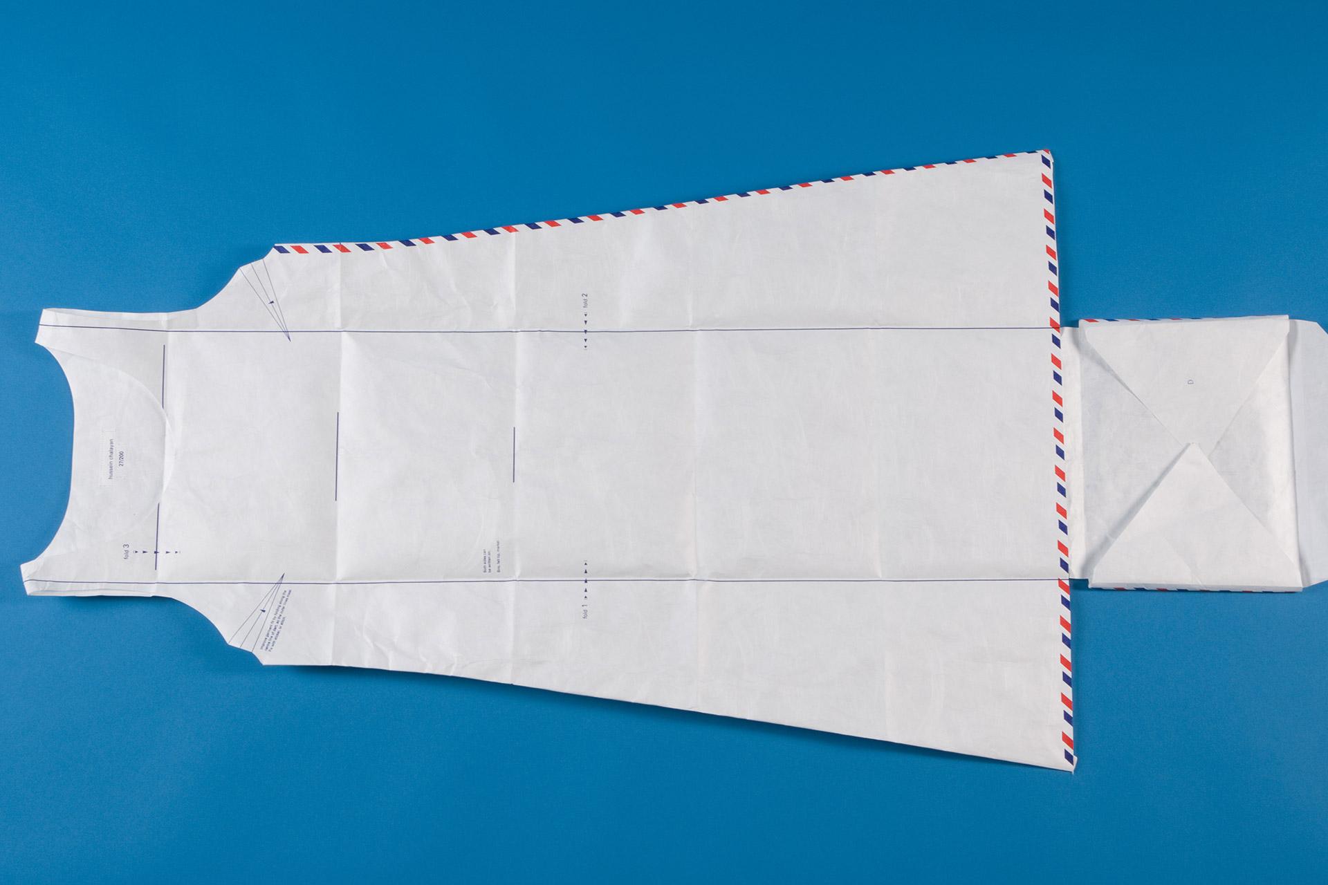 A dress pattern made from white paper with red and blue edging lies flat on a blue background. The pattern includes armholes, neckline, and folded edges, resembling the shape of a sleeveless dress.