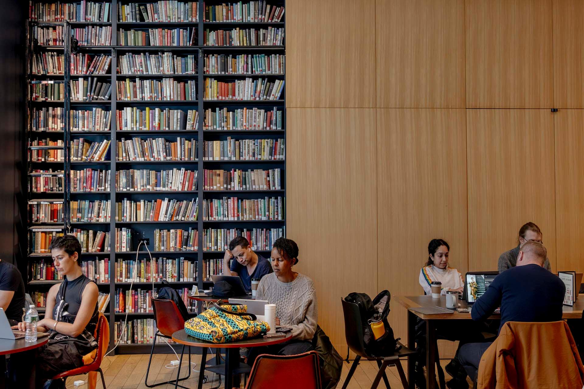 People studying and working on laptops at tables in a library. The room features a large wall filled with bookshelves and another wall with wooden paneling. Some individuals are reading while others are focused on their screens.