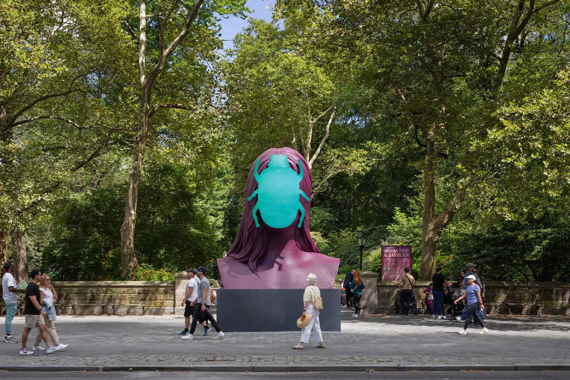 Several people walk past a large, colorful sculpture of a head wearing a turquoise face mask, set on a city sidewalk surrounded by tall trees and greenery.