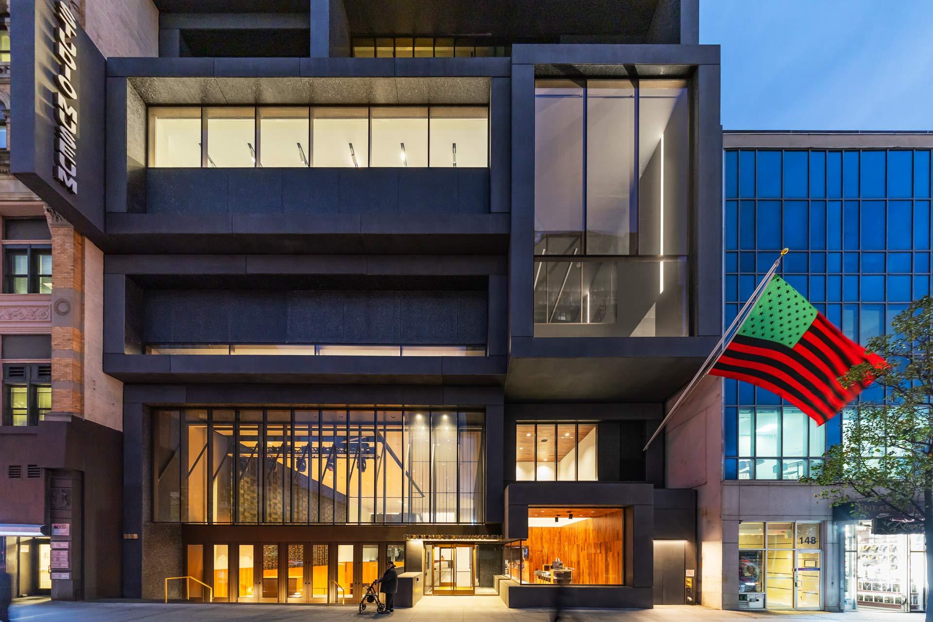 A modern building with large glass windows and dark geometric structures is shown at dusk. An American flag in red, black, and green hangs from the facade. A person pushes a stroller outside the entrance.