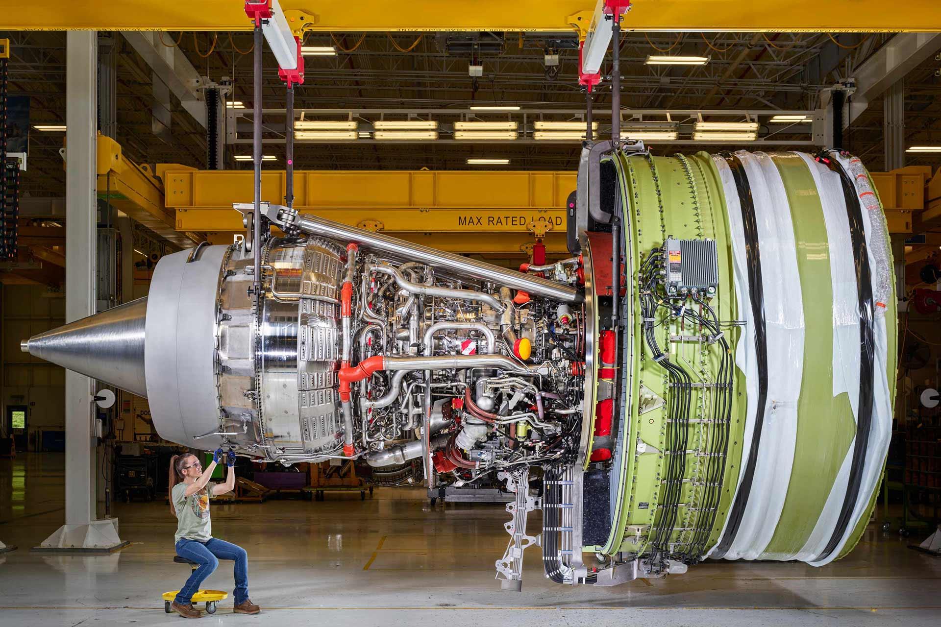 A person in safety gear inspects a large, exposed jet engine suspended in an industrial facility. The engine’s complex inner components and wiring are visible, and the person appears small beside the machinery.
