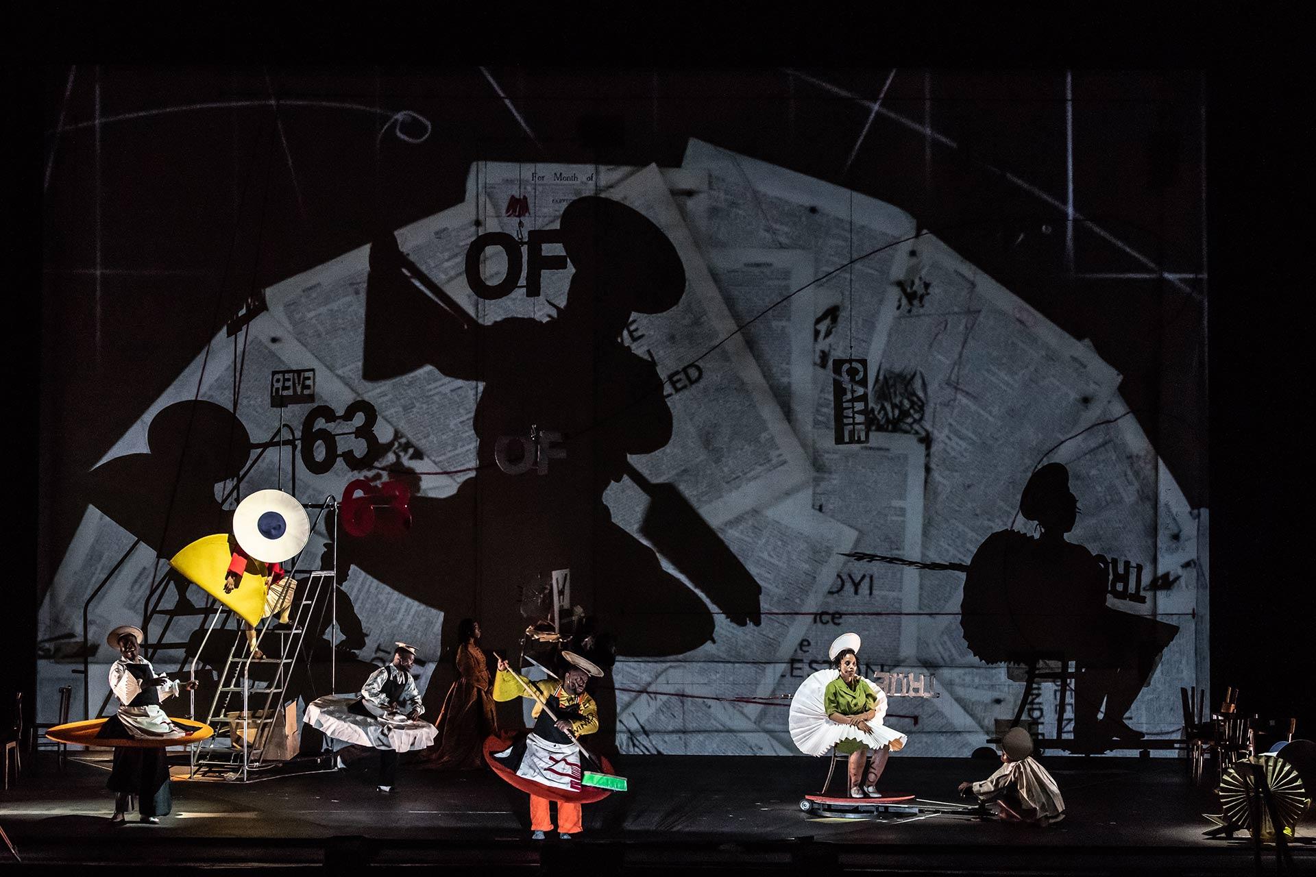 A surreal stage scene features performers in abstract, colorful costumes posed against a backdrop of newspaper clippings and bold shadows, creating an avant-garde, theatrical atmosphere.