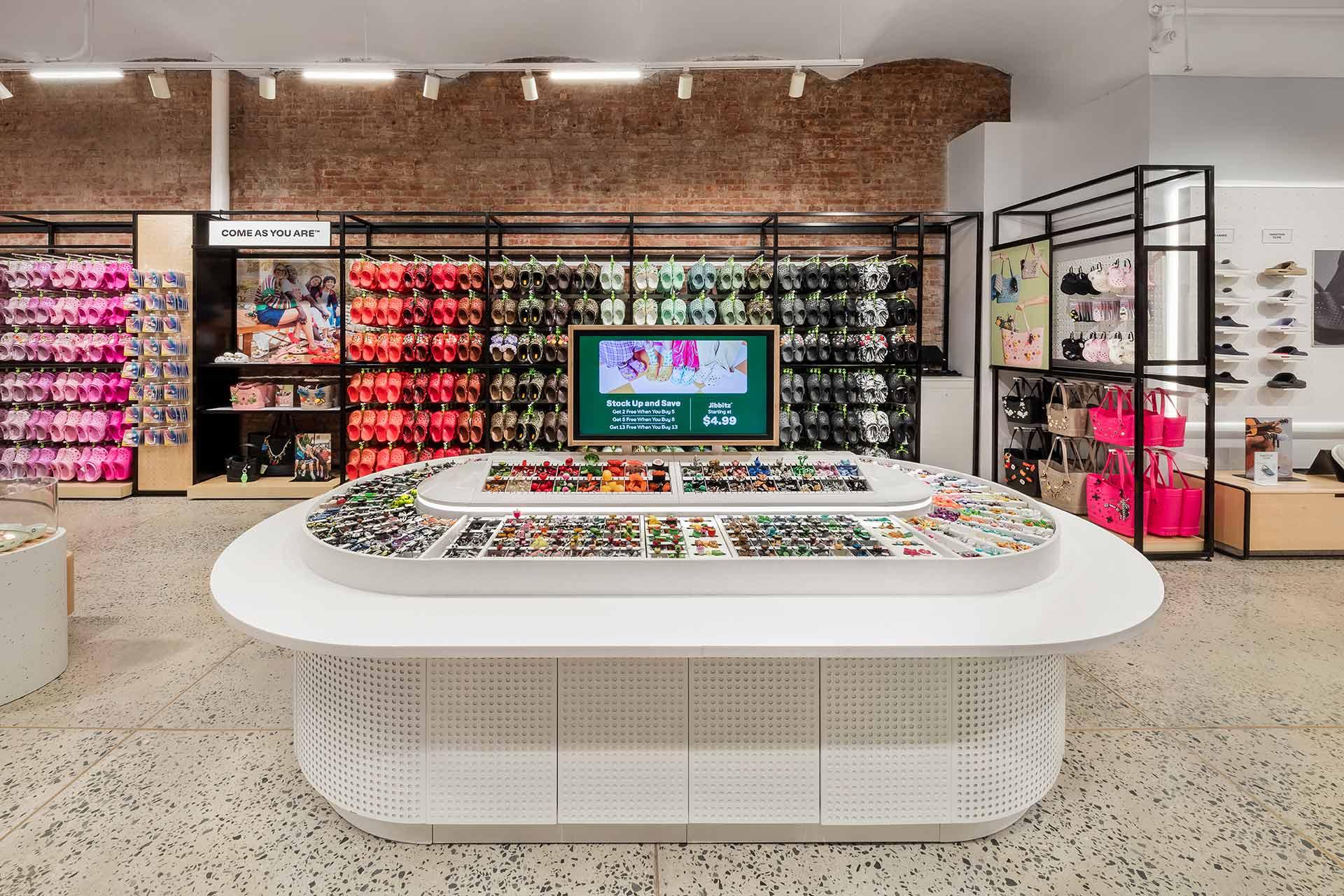 A brightly lit retail store with colorful footwear and bags displayed on wall racks, a central white display table filled with accessories, and a digital screen showcasing promotions. Brick and white walls create a modern aesthetic.