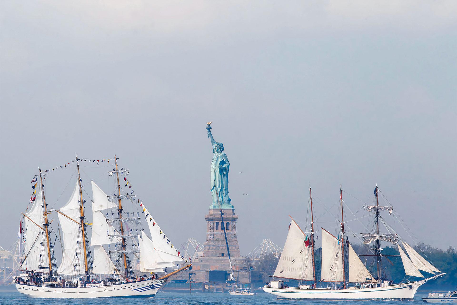 Tall sailing ships with white sails pass by the Statue of Liberty on a hazy day. The statue stands on its pedestal, and the ships are decorated with colorful flags as they sail on the water.