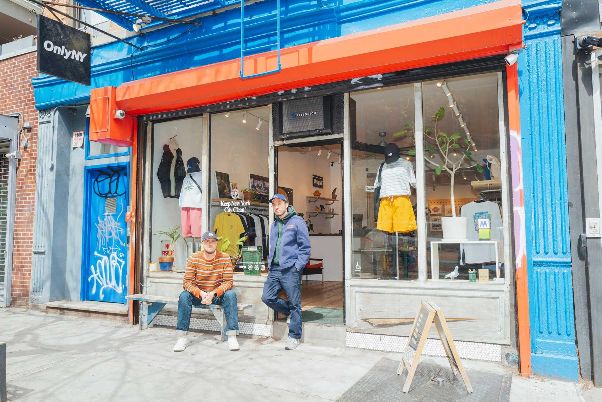 Julian and Micah wearing casual clothes stand in front of the OnlyNY storefront with a glass display window, mannequins, and street art graffiti on the adjacent wall.