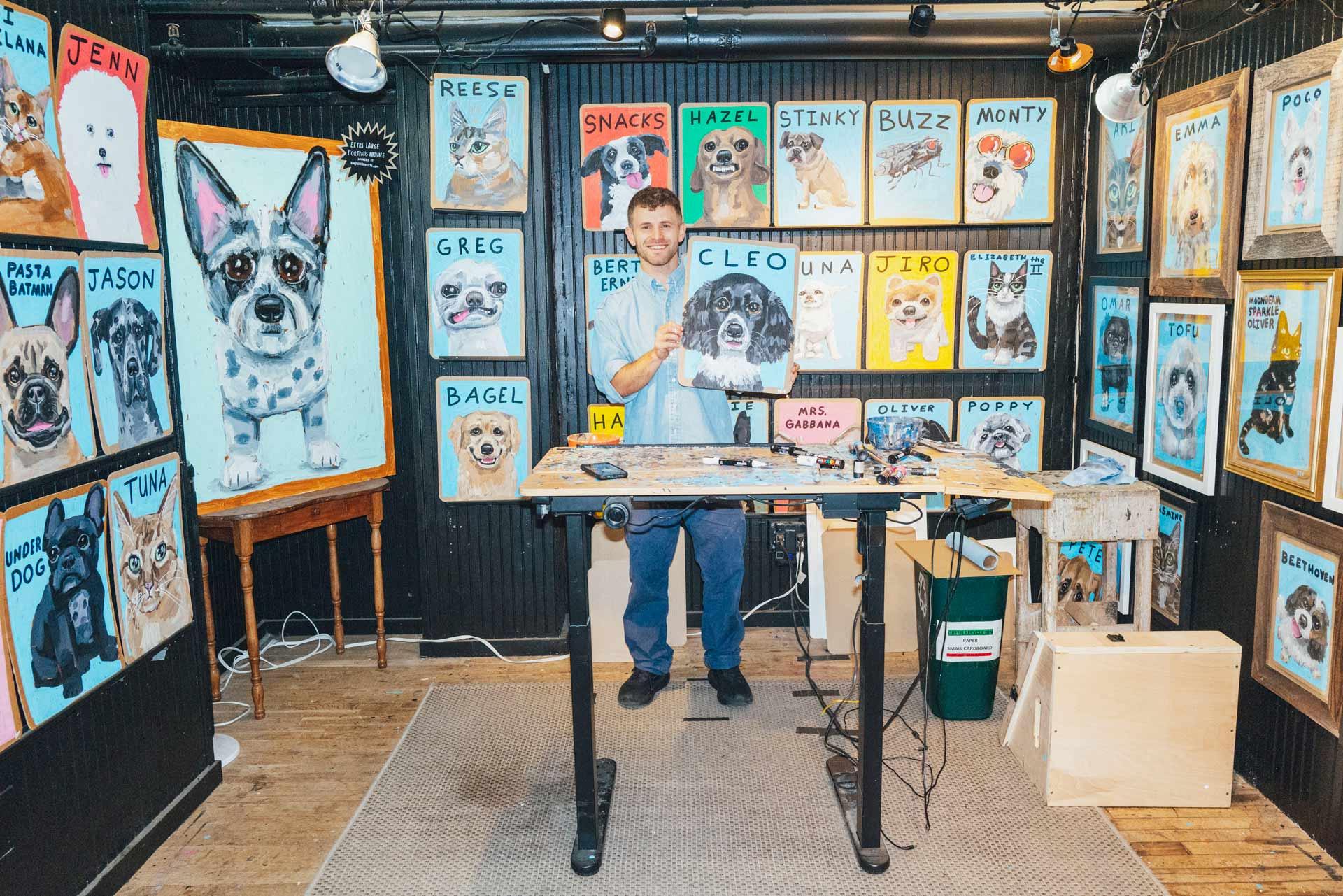 Ben Lenovitz stands in his studio holding a dog portrait. The walls behind him are covered with colorful, framed dog paintings, each labeled with a different name. Art supplies are on a desk in front of him.