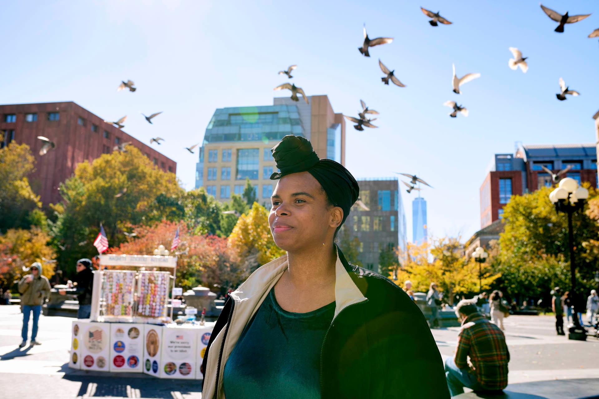 A woman with a green headwrap stands outdoors in a city park on a sunny day, with pigeons flying overhead and people in the background among autumn-colored trees and urban buildings.