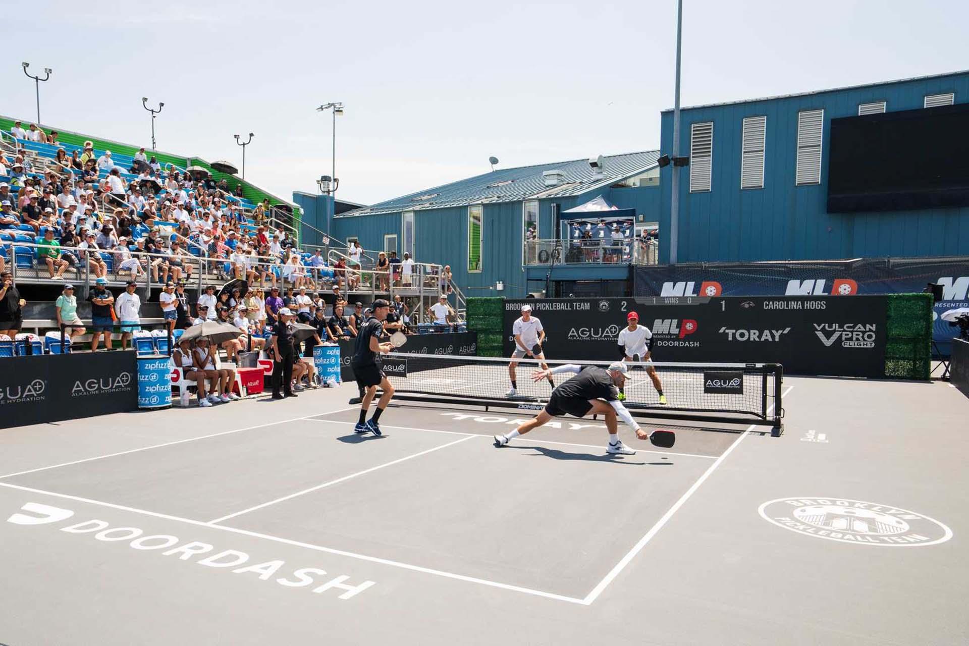 Four athletes play a competitive pickleball doubles match on an outdoor court, with a large crowd of spectators watching from bleachers under a sunny sky. Sponsor logos are visible around the court.