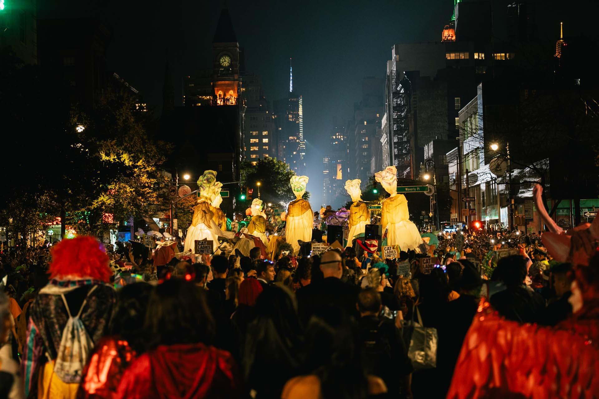 A large crowd gathers at night for the Halloween Parade, with people in colorful costumes and several large illuminated animal head masks held above the crowd. City buildings and street signs are visible in the background.