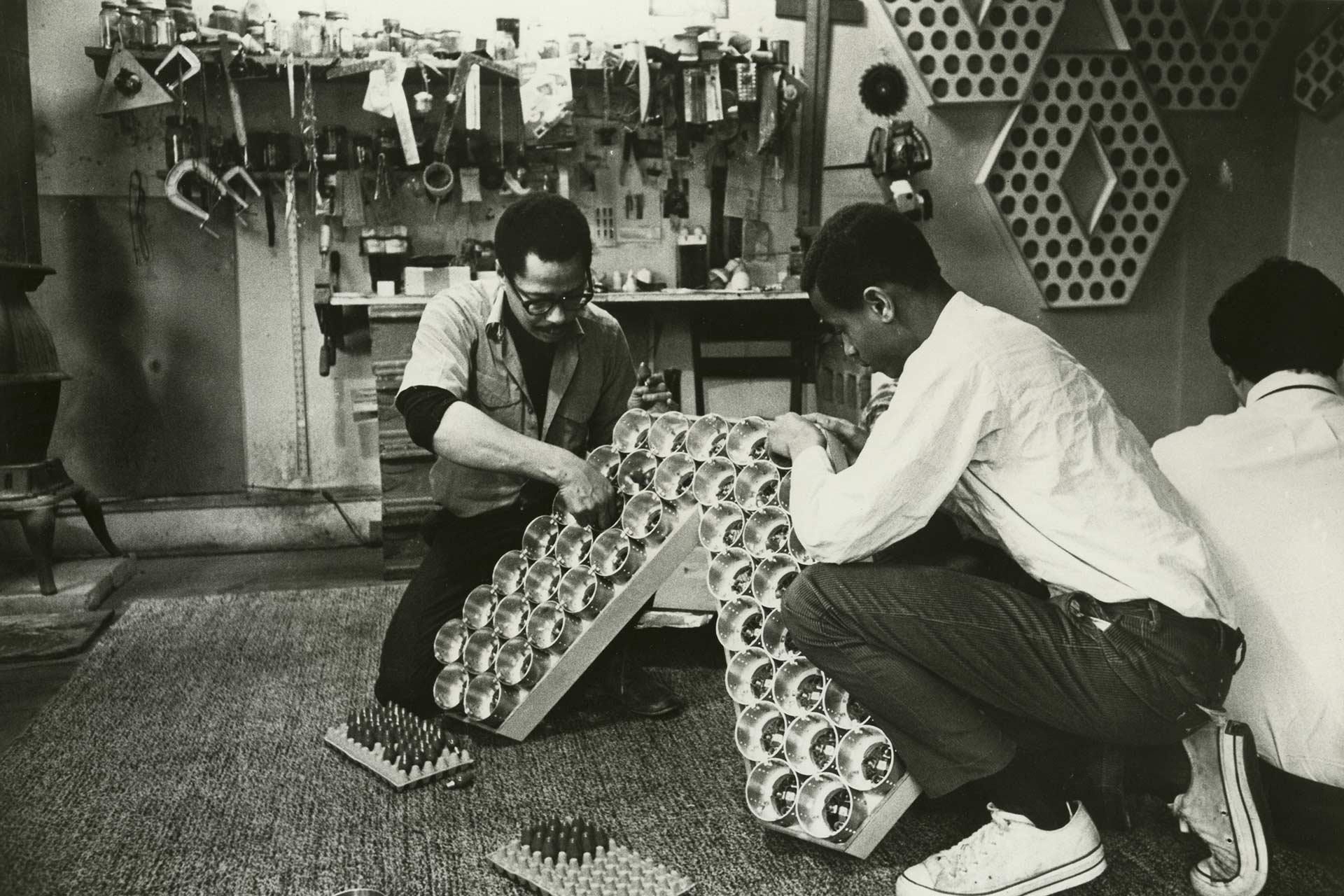 Two people kneel on the floor, assembling a structure made of large clear bulbs in a workshop filled with tools and pegboards. Another person sits nearby with their back to the camera.