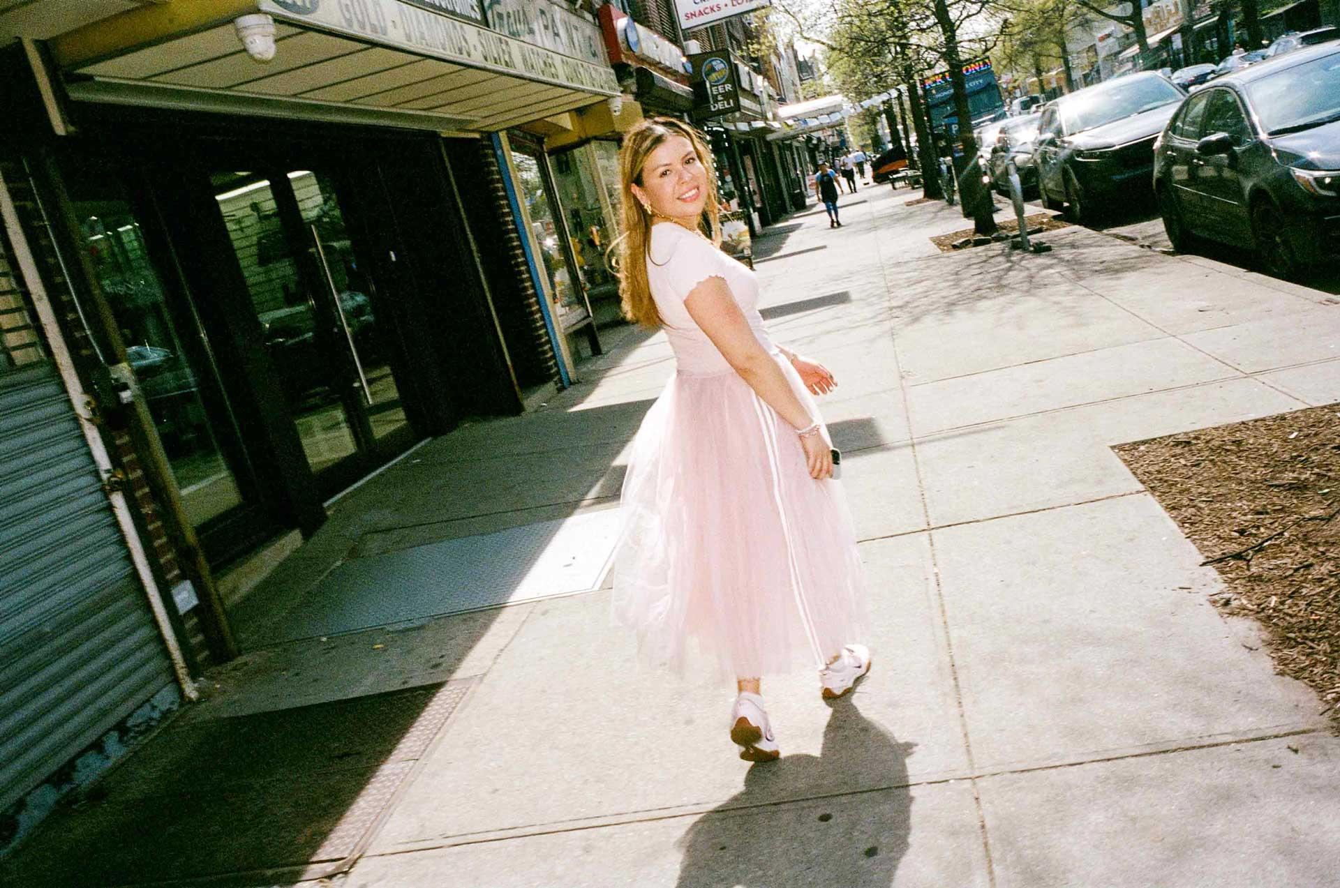Maria-Isabelle in a light pink dress smiles and looks back while walking on a sunny city sidewalk lined with shops and parked cars.