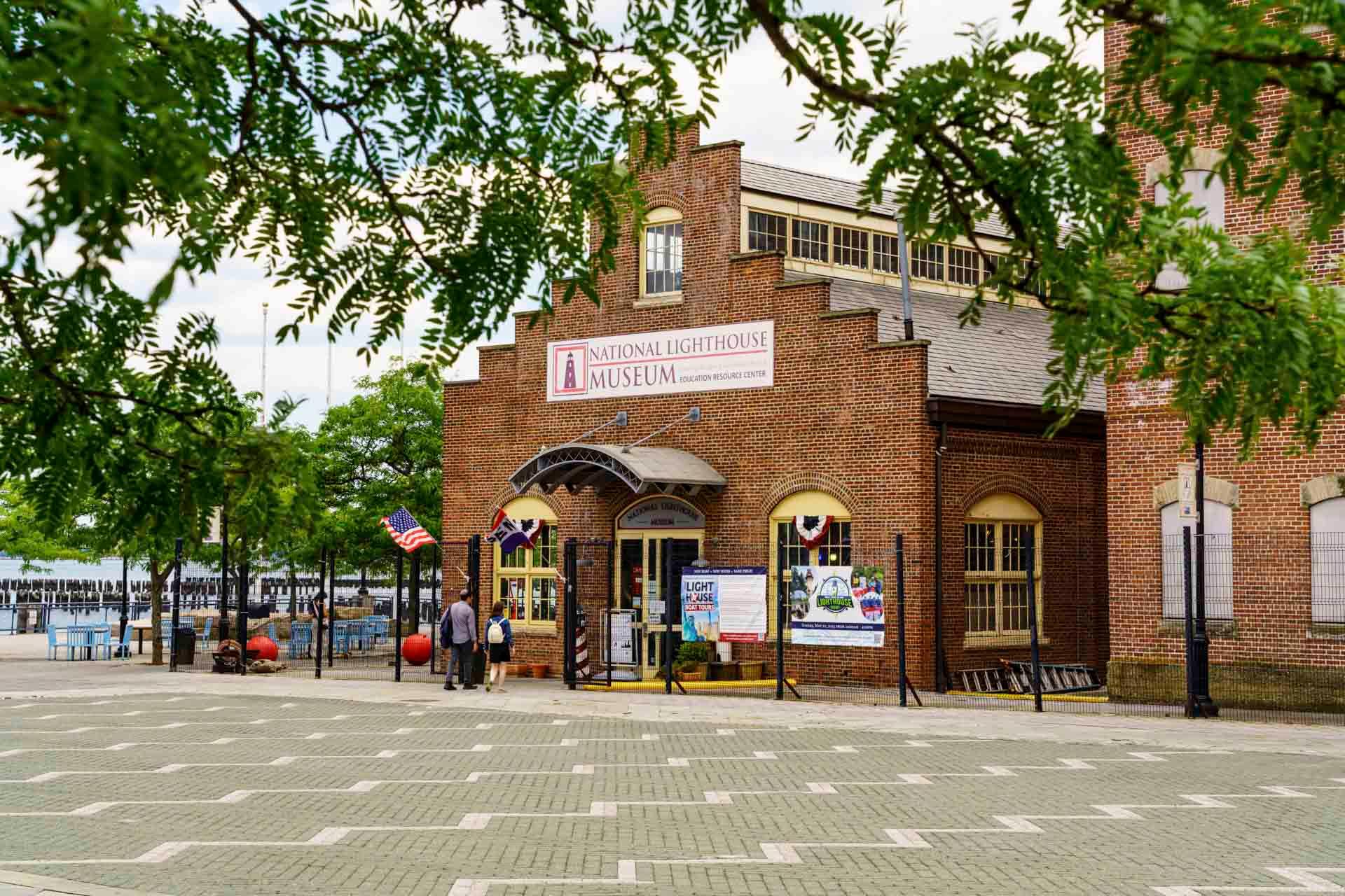 The image shows the National Lighthouse Museum, a red brick building with arched windows and a sign above the entrance. There are informational posters, American flags, and trees framing the scene. People are walking towards the entrance.