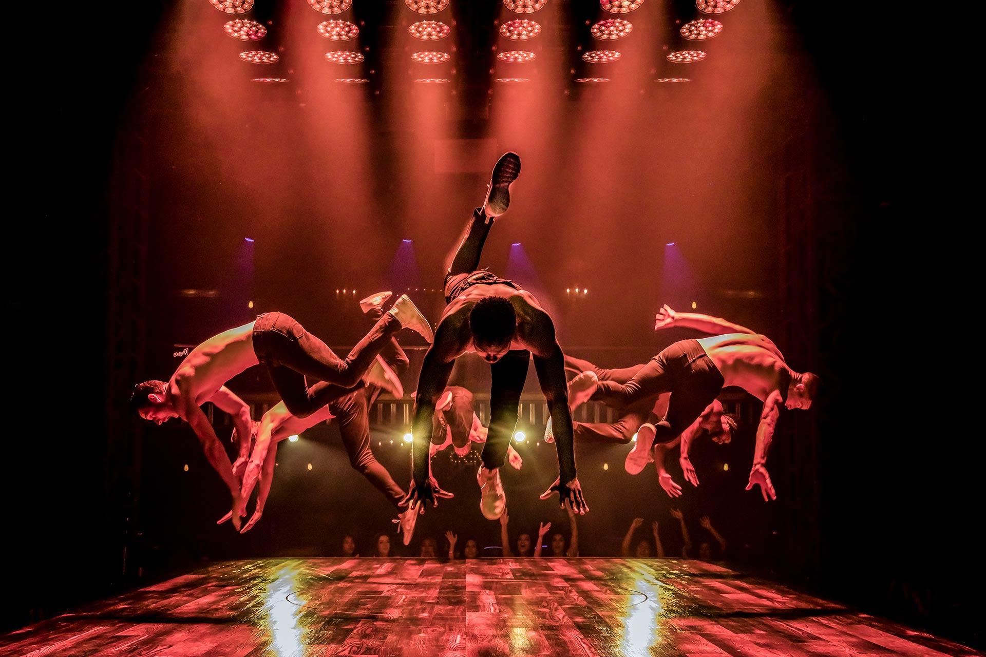 Five dancers perform a synchronized jump on stage under dramatic red lighting, captured mid-air with dynamic poses and reflections on the shiny floor, creating an energetic and powerful scene.