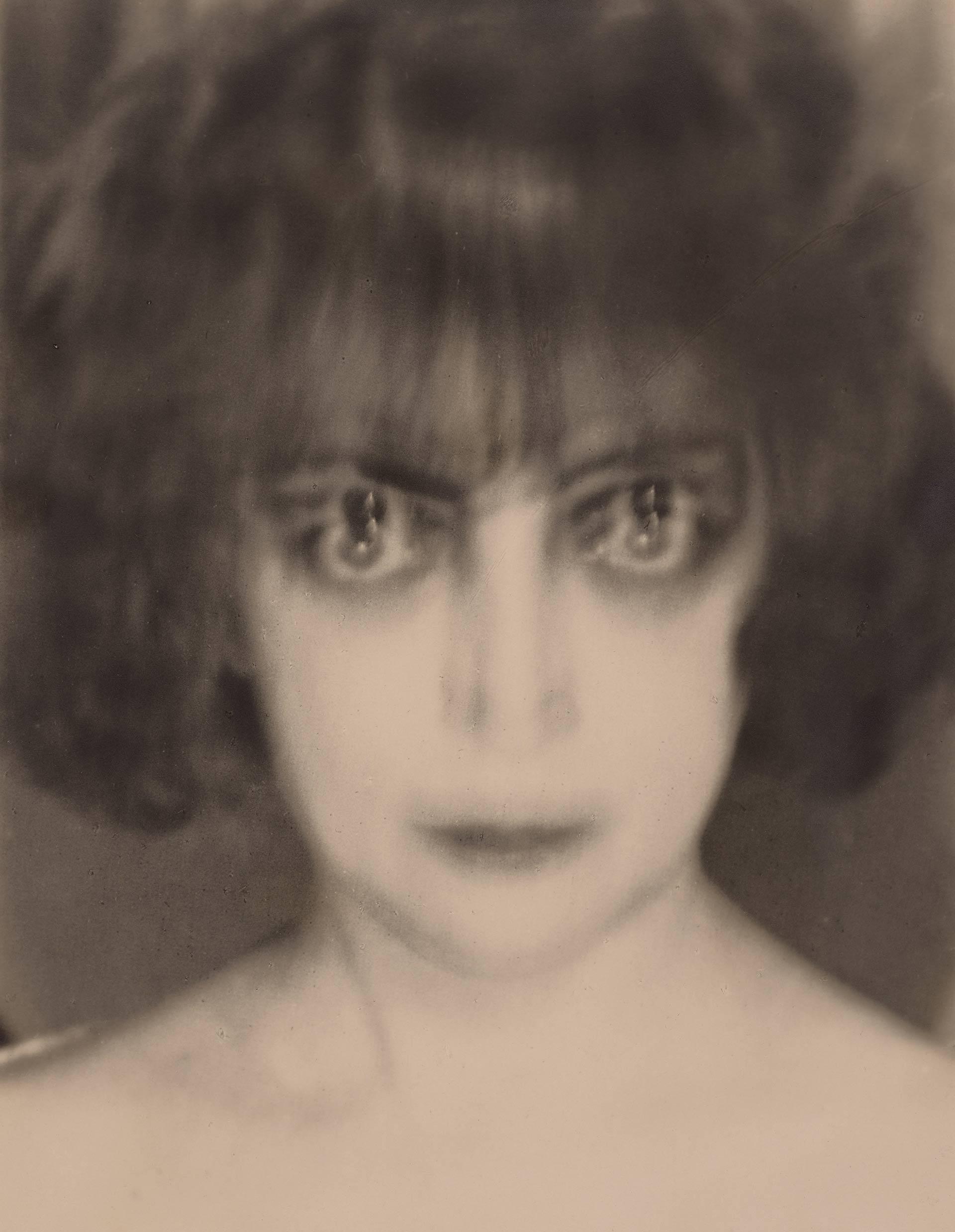 A blurred, black-and-white portrait of a person with short, wavy hair and large, darkly emphasized eyes staring directly at the camera, creating a surreal and intense effect.