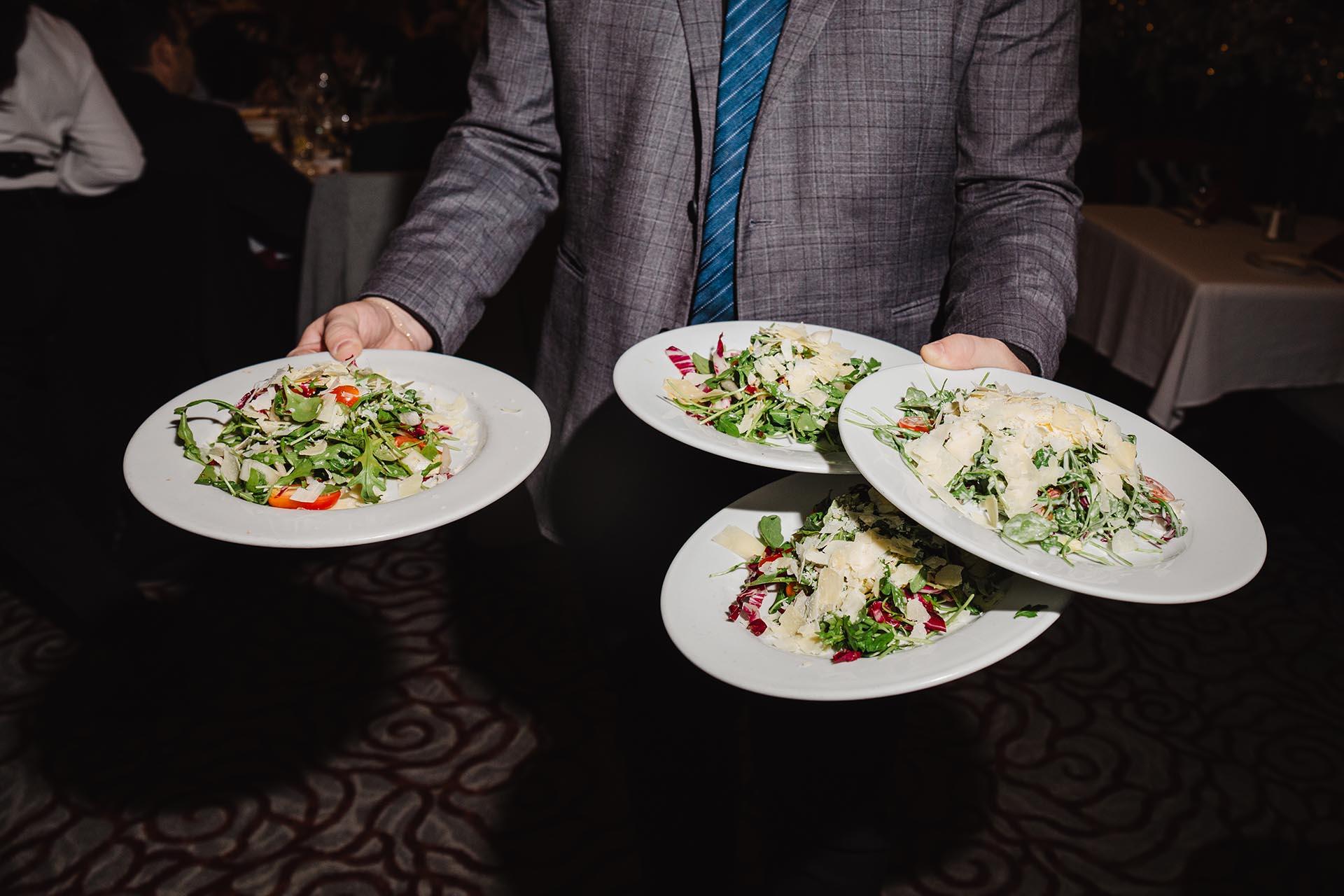 A person in a suit and blue striped tie carries four white plates of salad and pasta dishes, balanced skillfully in both hands, in a dimly lit restaurant setting.