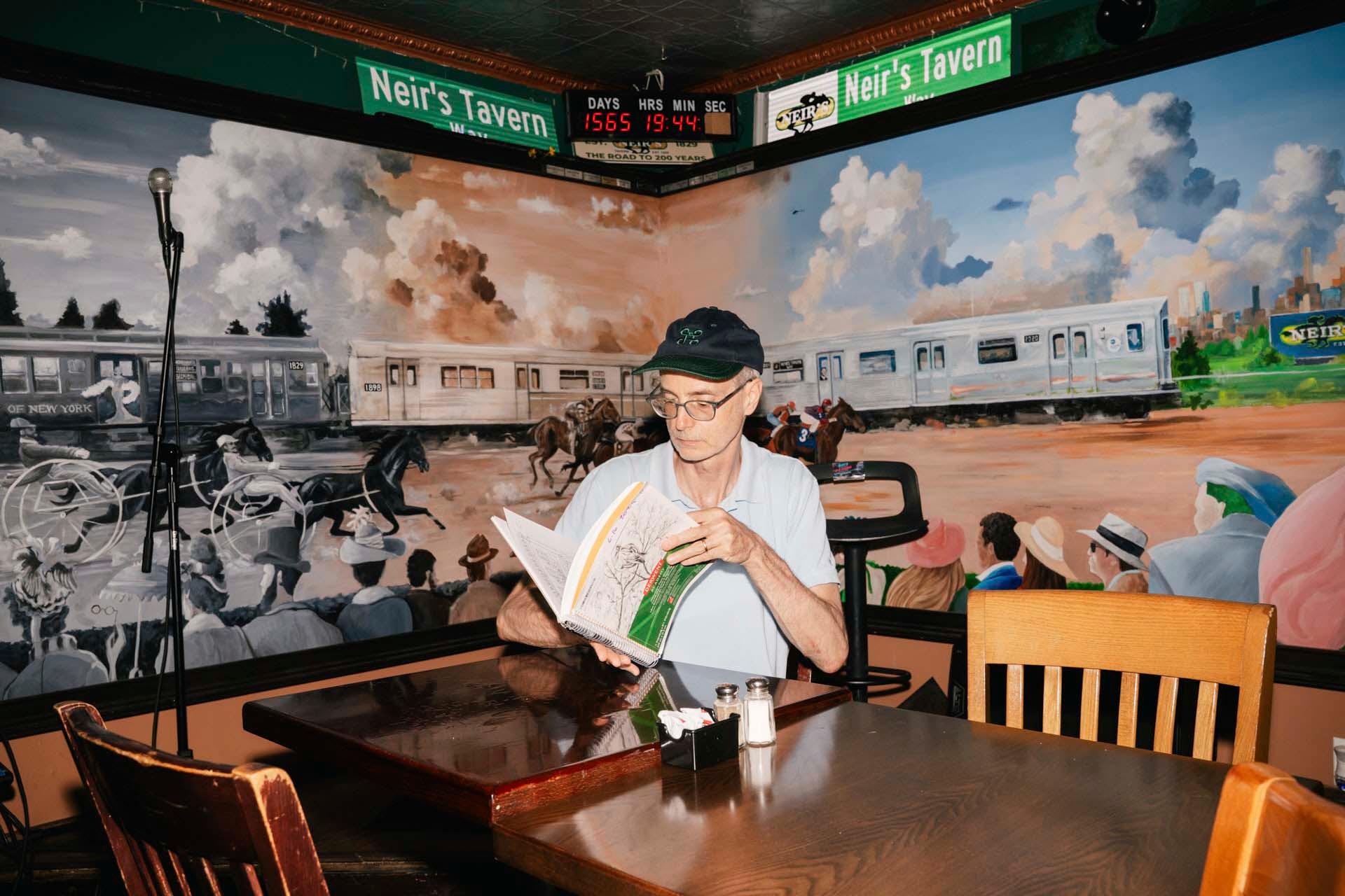 A man wearing glasses and a cap reads a book at a wooden table in Neir’s Tavern, with a large mural of a horse race and subway train covering the wall behind him. An empty microphone stands nearby.