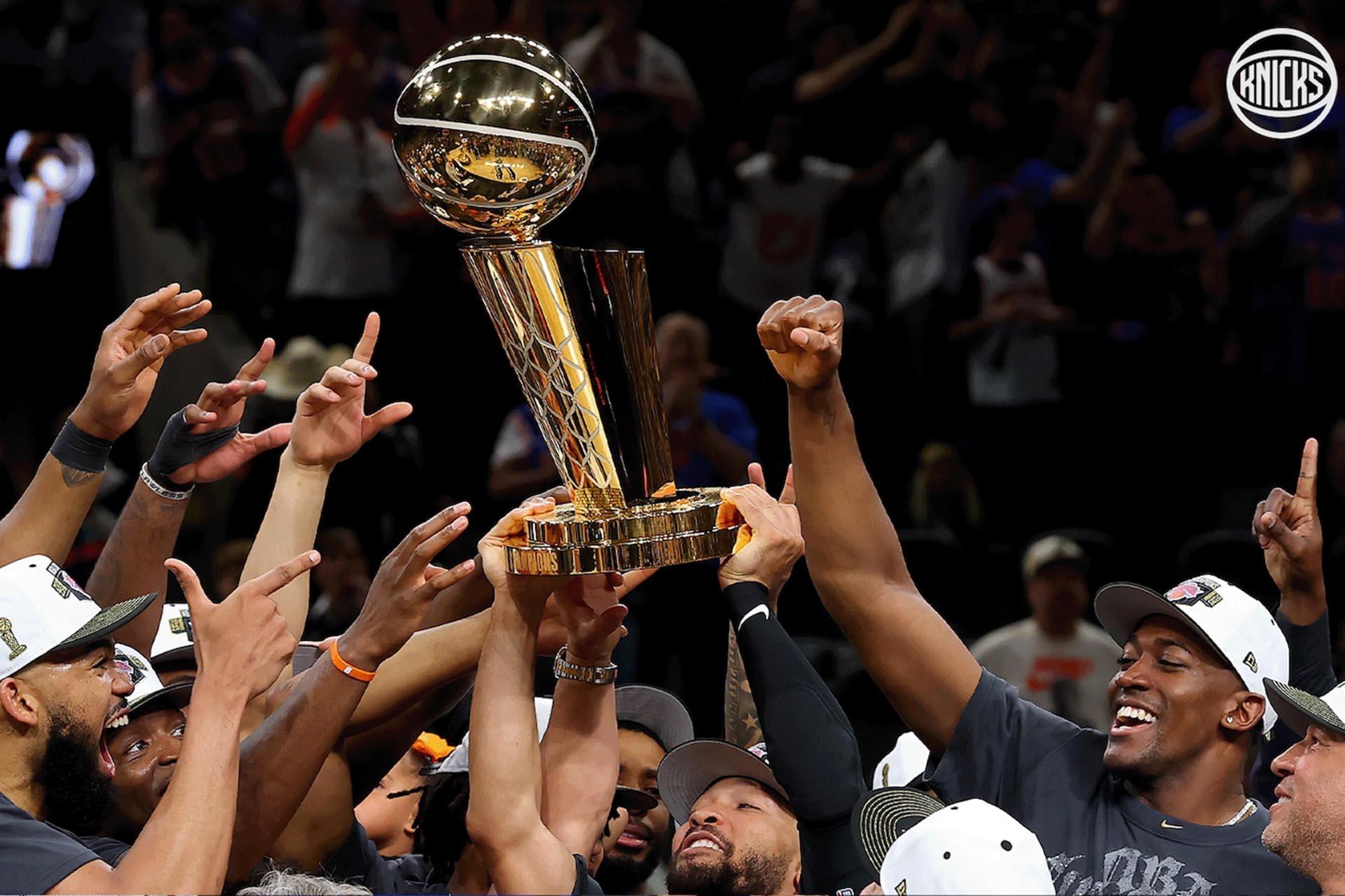 Basketball players in black shirts and caps celebrate by raising a golden NBA championship trophy together, surrounded by cheering teammates and fans. The Knicks logo is visible in the top right corner.
