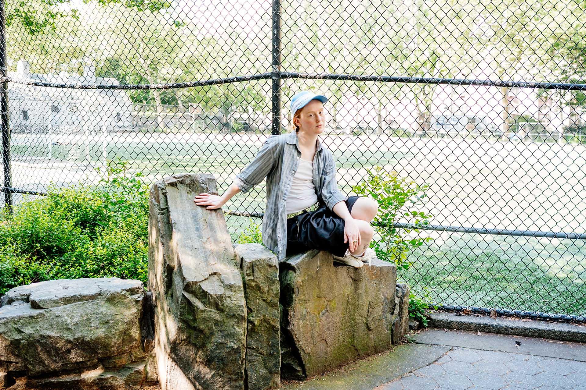 A person wearing a blue cap, striped shirt, white t-shirt, and shorts sits relaxed on stone steps outside a white columned building surrounded by greenery.