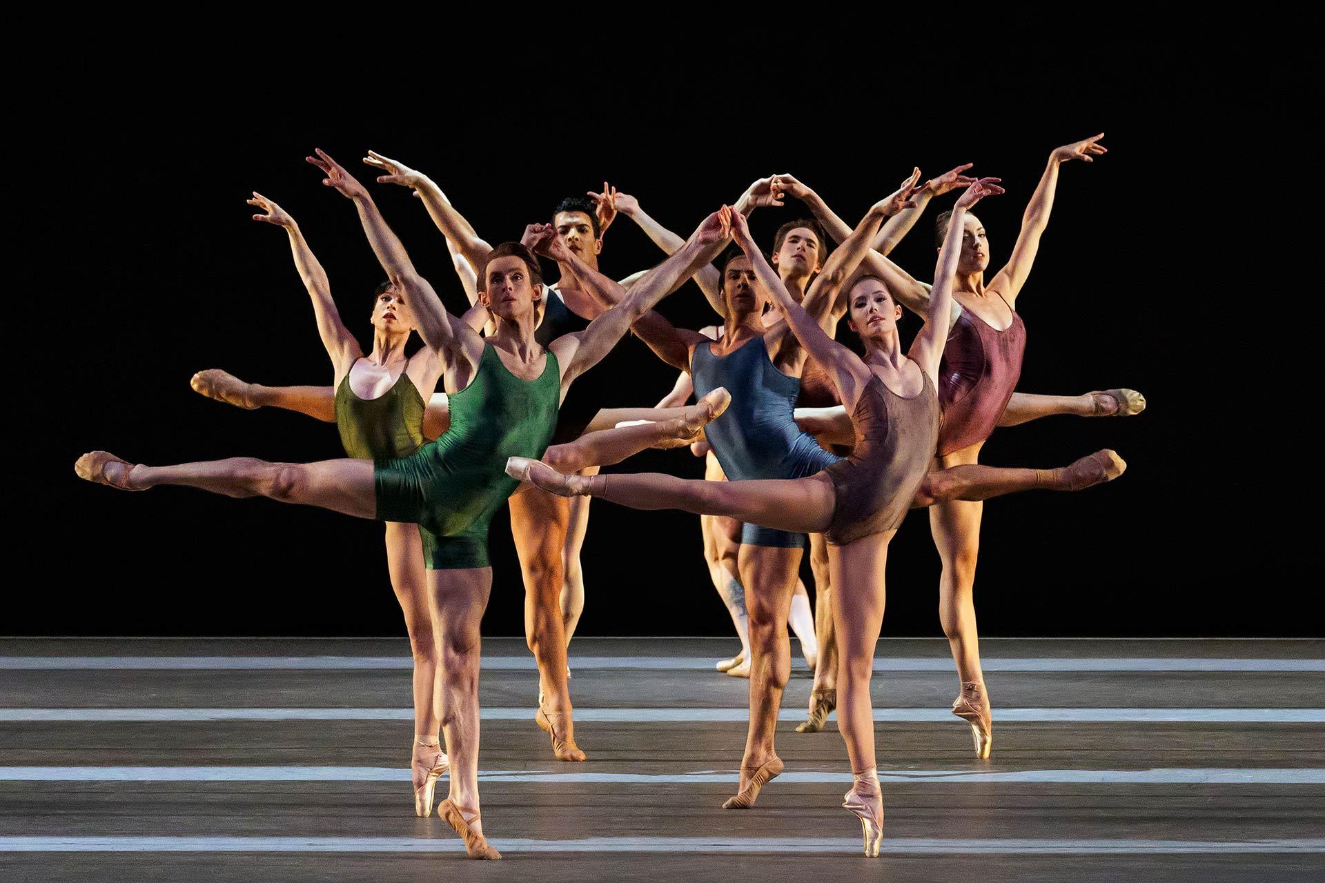Eight ballet dancers in colorful leotards perform on stage, extending one leg and both arms gracefully in unison, creating a symmetrical and dynamic formation against a dark background.