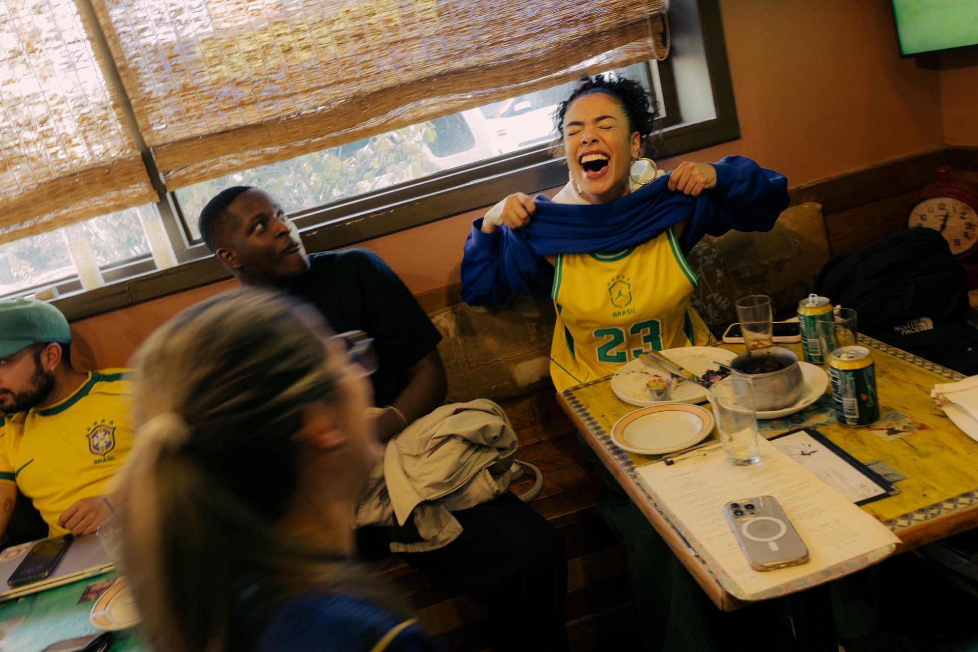 A woman joyfully lifts her Brazil jersey while sitting at a table in a lively restaurant, surrounded by friends, empty plates, and drinks, creating a festive and energetic atmosphere.