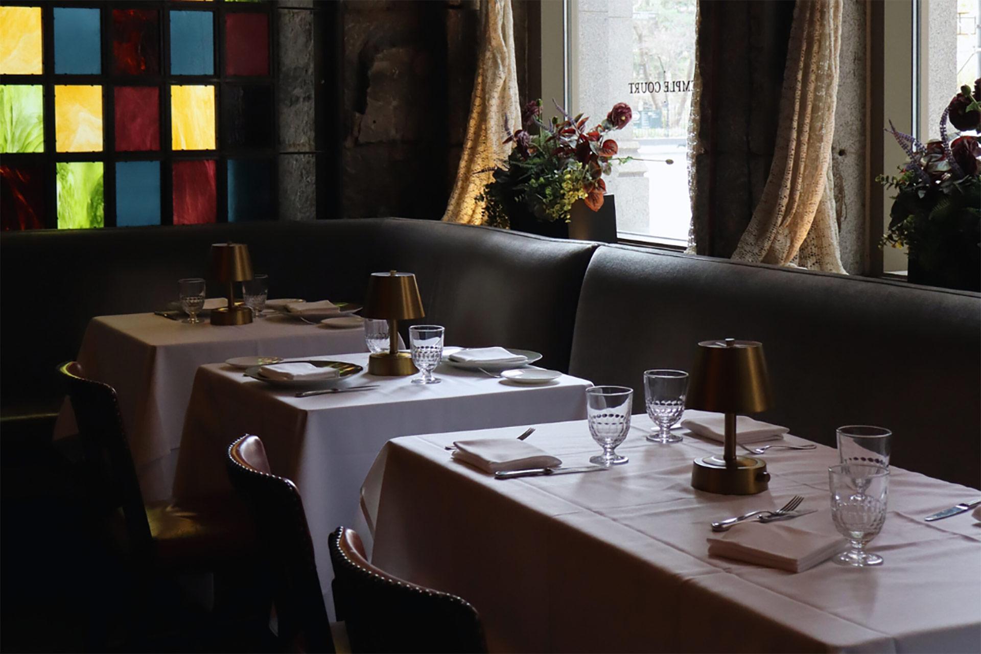 Elegant restaurant interior with tables set for two, each with white tablecloths, glassware, napkins, and small brass lamps; stained glass window and large curtains in the background with floral arrangements.