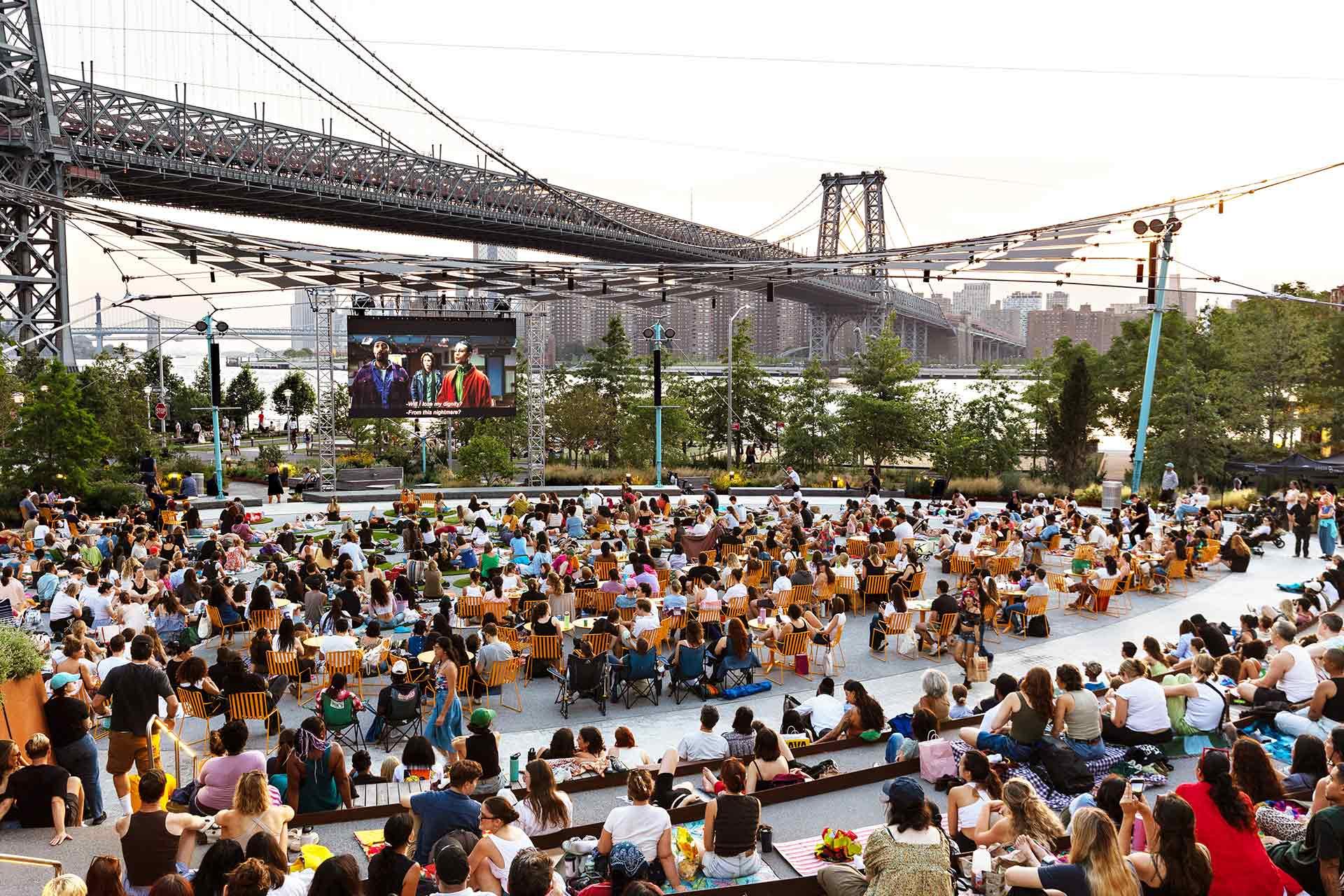 A large crowd gathers outdoors under a bridge to watch a movie on a big screen, with chairs and blankets spread out on the ground and city buildings visible in the background.