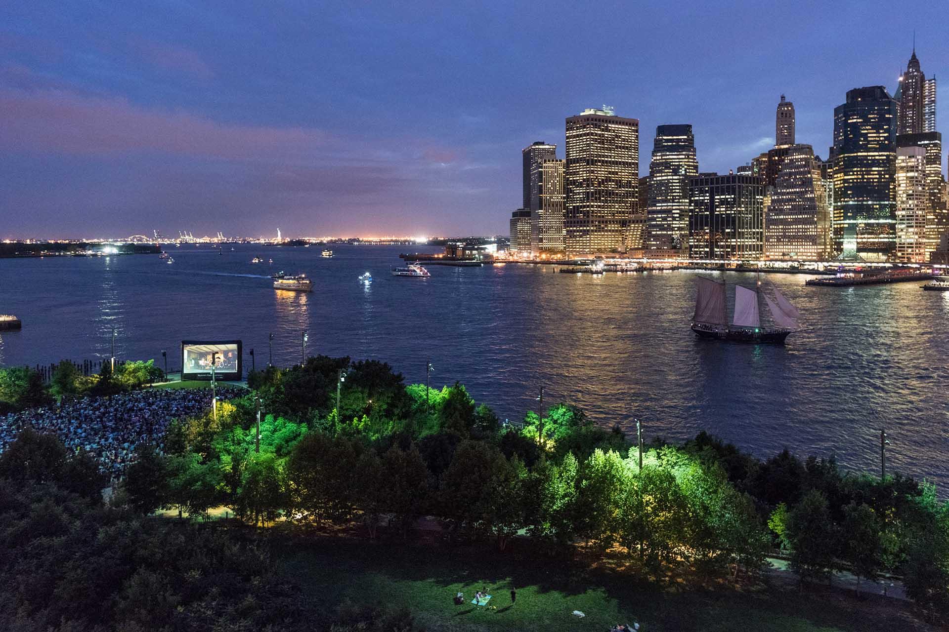 An outdoor movie screening takes place in a park by the water at dusk, with crowds gathered near a large screen. Across the river, a city skyline with illuminated skyscrapers is visible, and boats sail on the water.