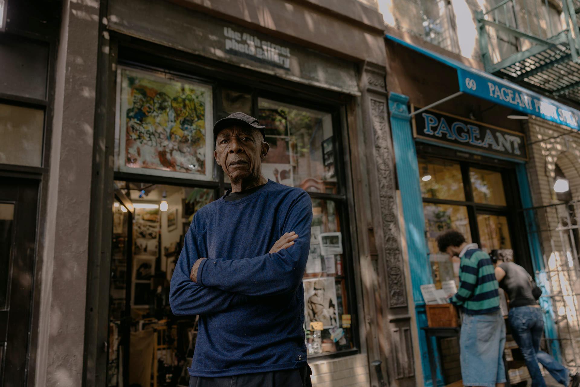 Alex Harsley in a dark blue sweater and cap stands with arms crossed in front of The 4th Street Photo Gallery.